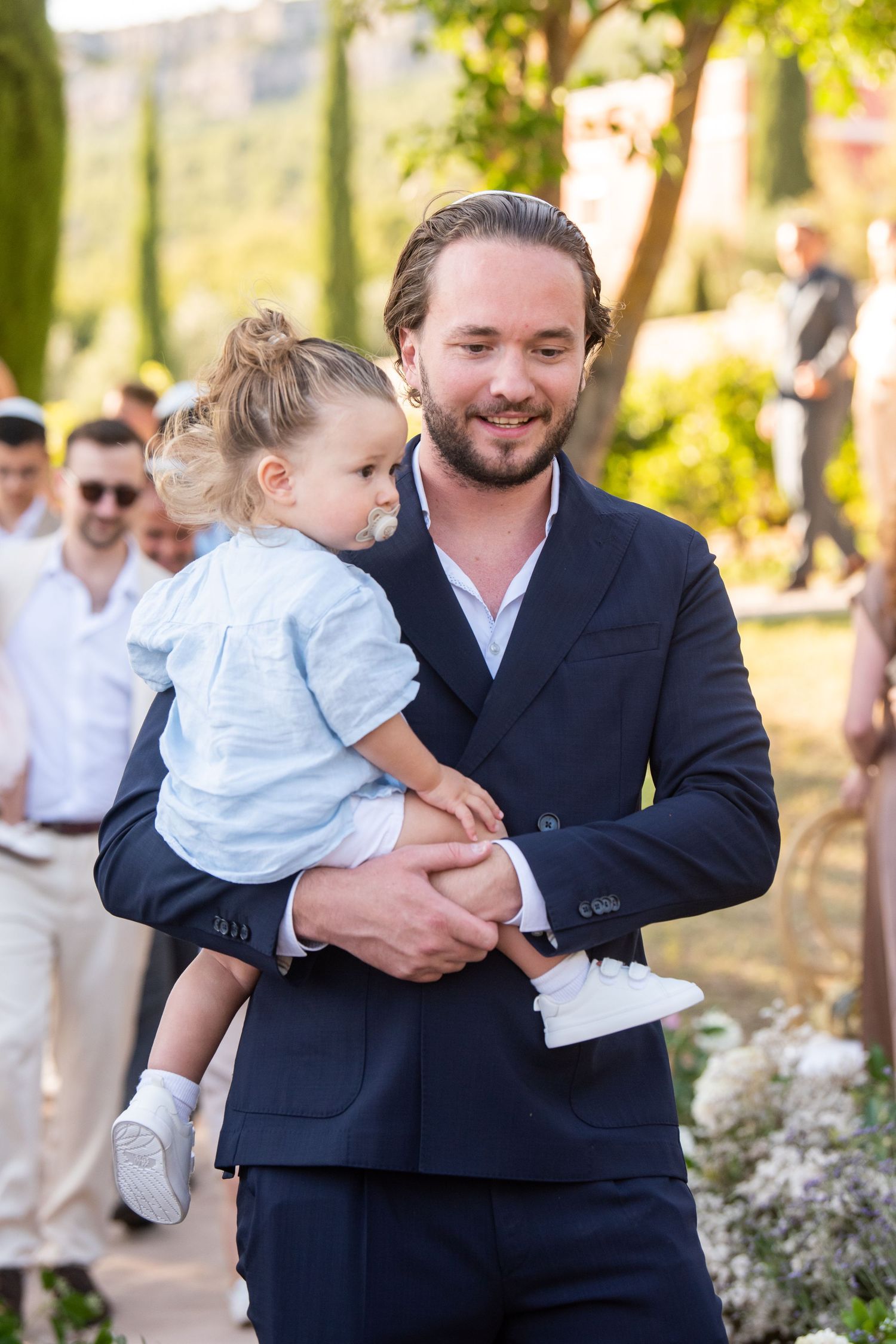 A man in a navy suit holds a small child wearing gray at an outdoor wedding ceremony with floral decorations.