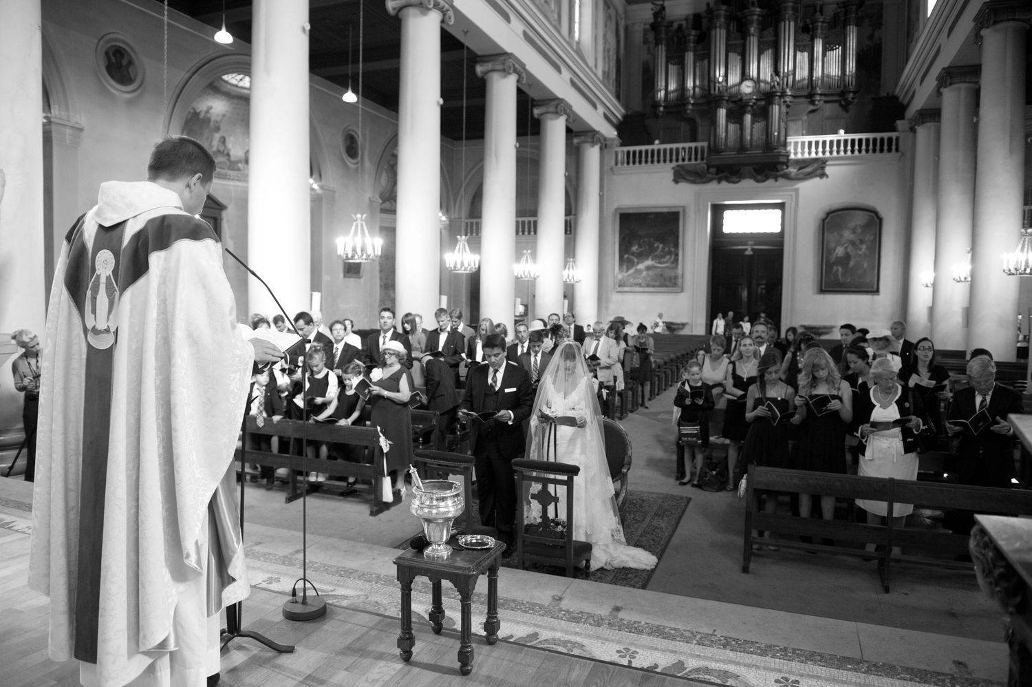 Black and white photo of a wedding ceremony in a church with guests seated in pews as the couple stands at the altar.