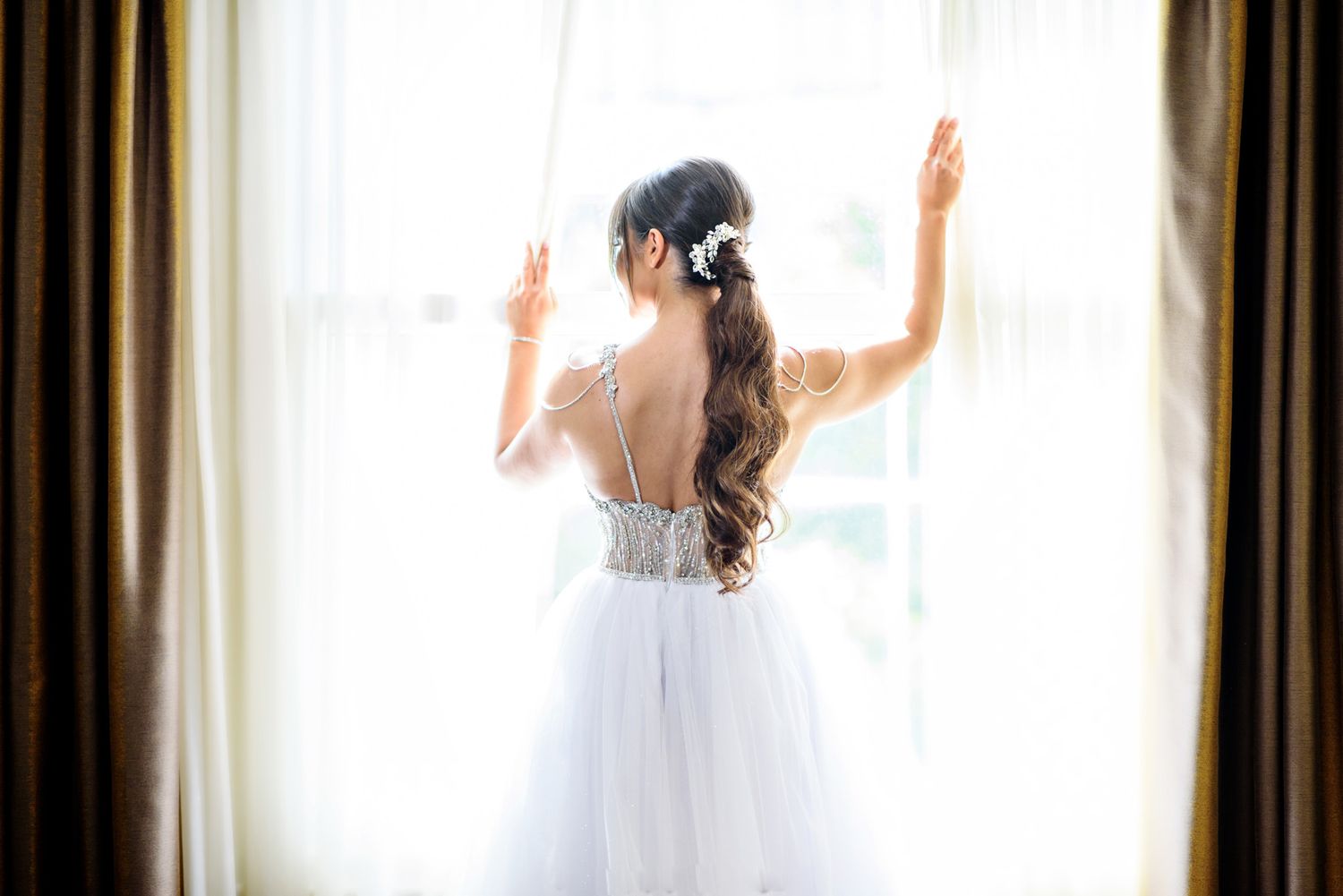 Bride in white tulle wedding dress with floral hair accessory stands facing window with arms raised.