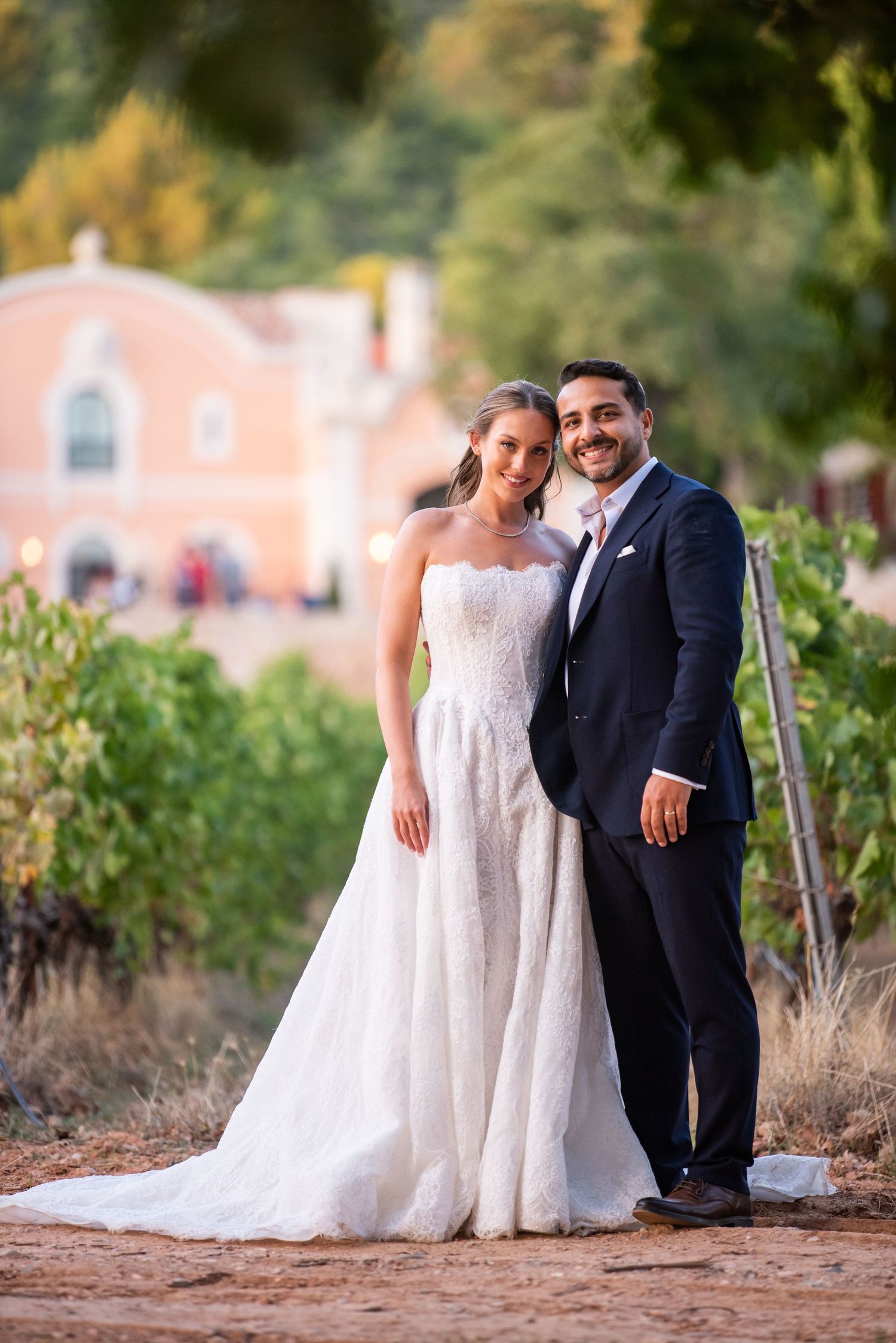 Bride in white gown and groom in navy suit pose together in vineyard with pink building backdrop.