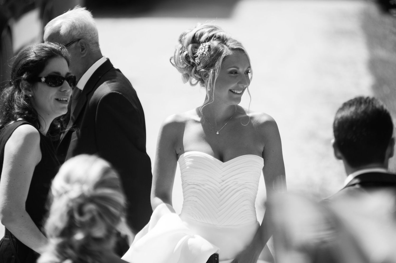 Smiling bride in strapless white gown walks with guests at outdoor wedding ceremony in black and white photo.