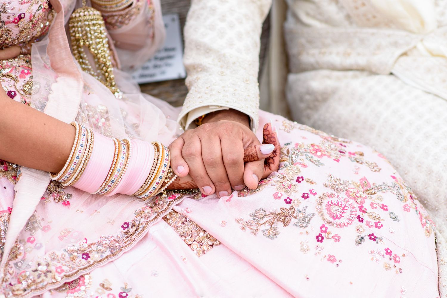 Indian bride and groom holding hands in traditional pink and white wedding attire adorned with gold bangles and embroidery.