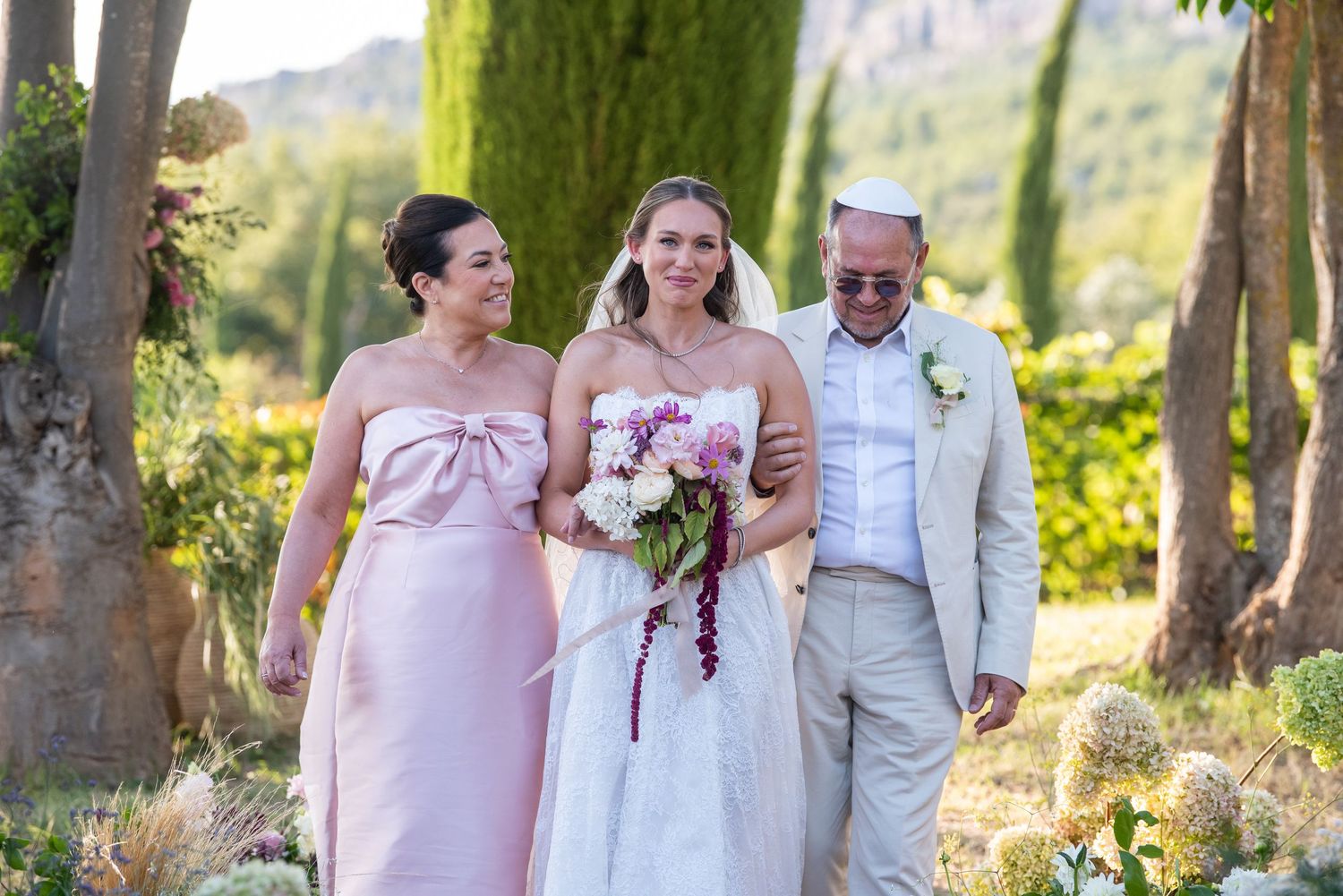 A group of people in formal attire pose together in a sunlit forest setting during a wedding celebration.