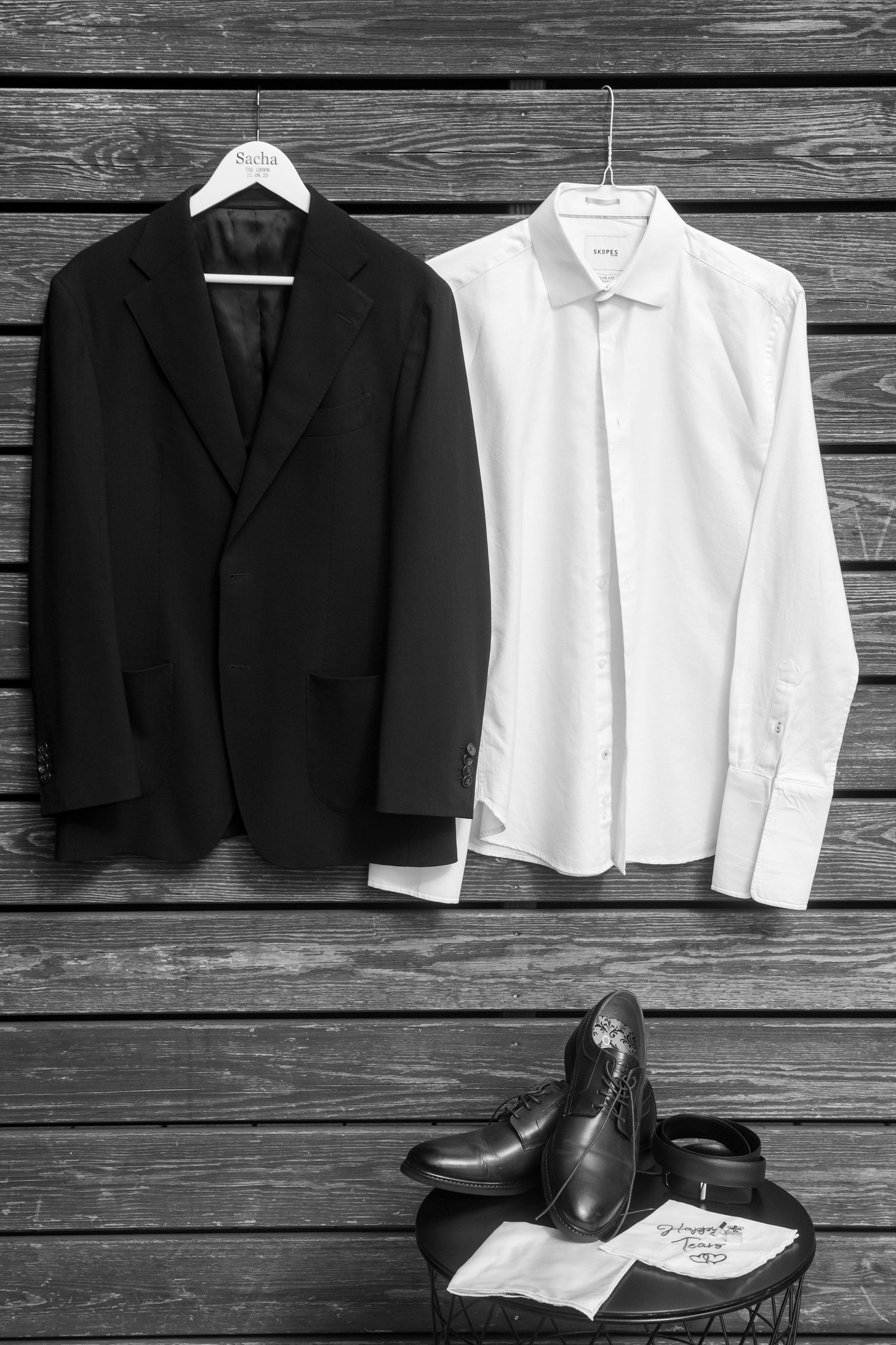 Black suit jacket and white dress shirt laid flat on rustic wooden planks with formal shoes below.
