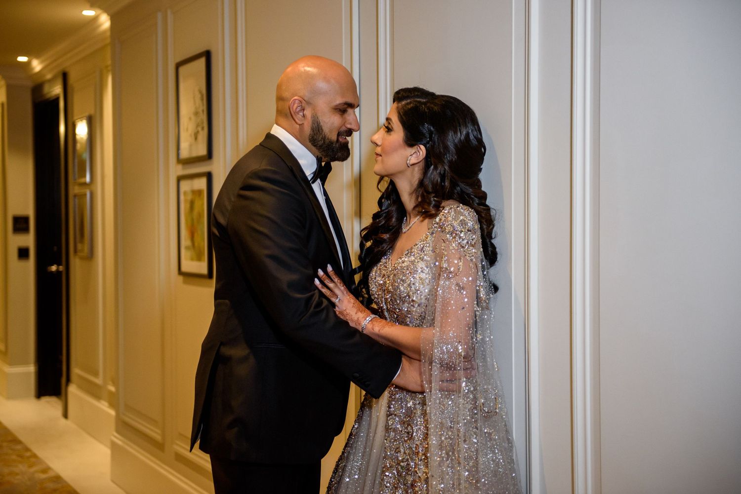 A couple in formal attire embraces in an elegant hallway with warm lighting and framed artwork on the walls.
