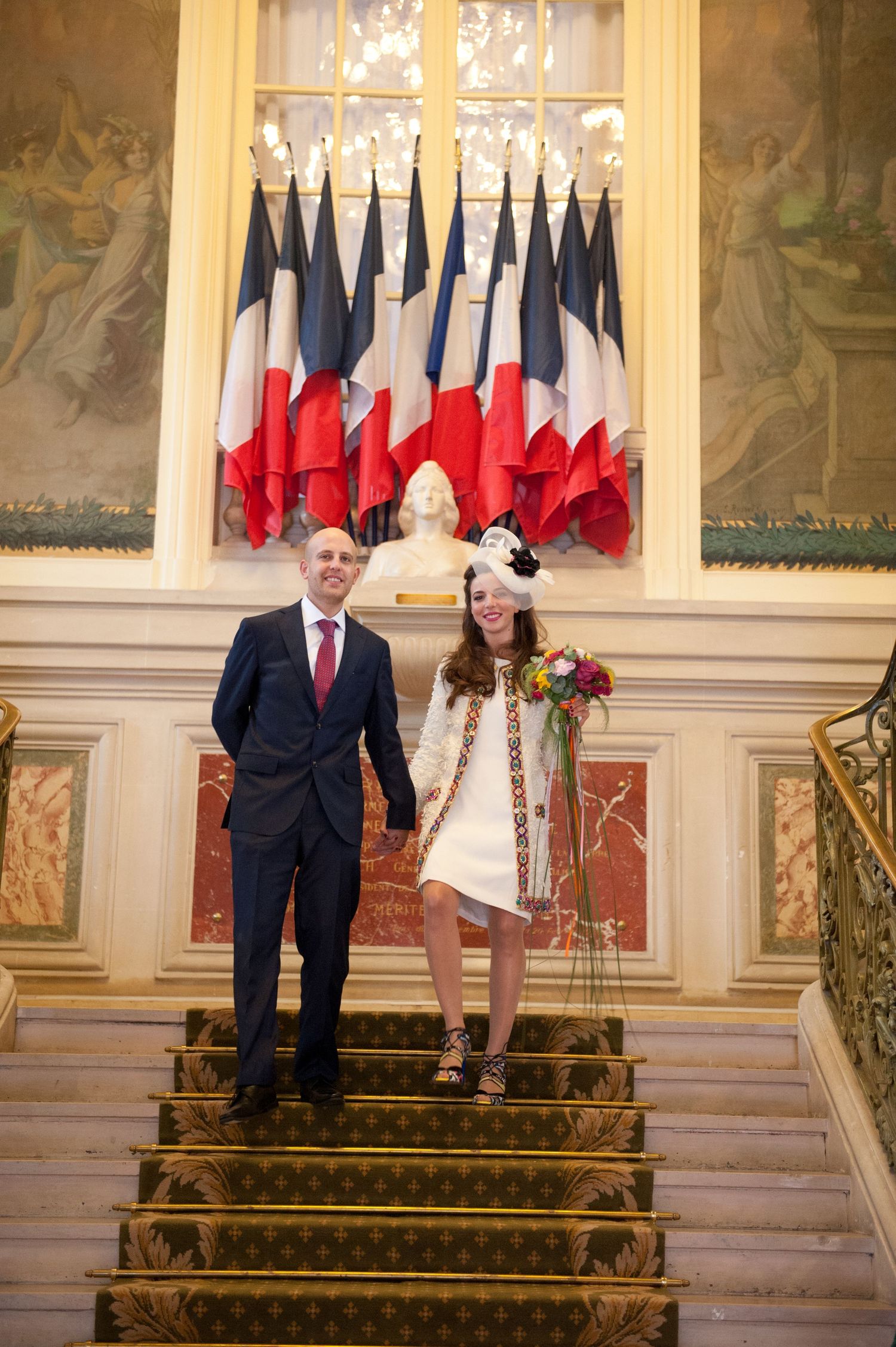 A couple descends an ornate staircase in a formal setting with French flags displayed in the background.