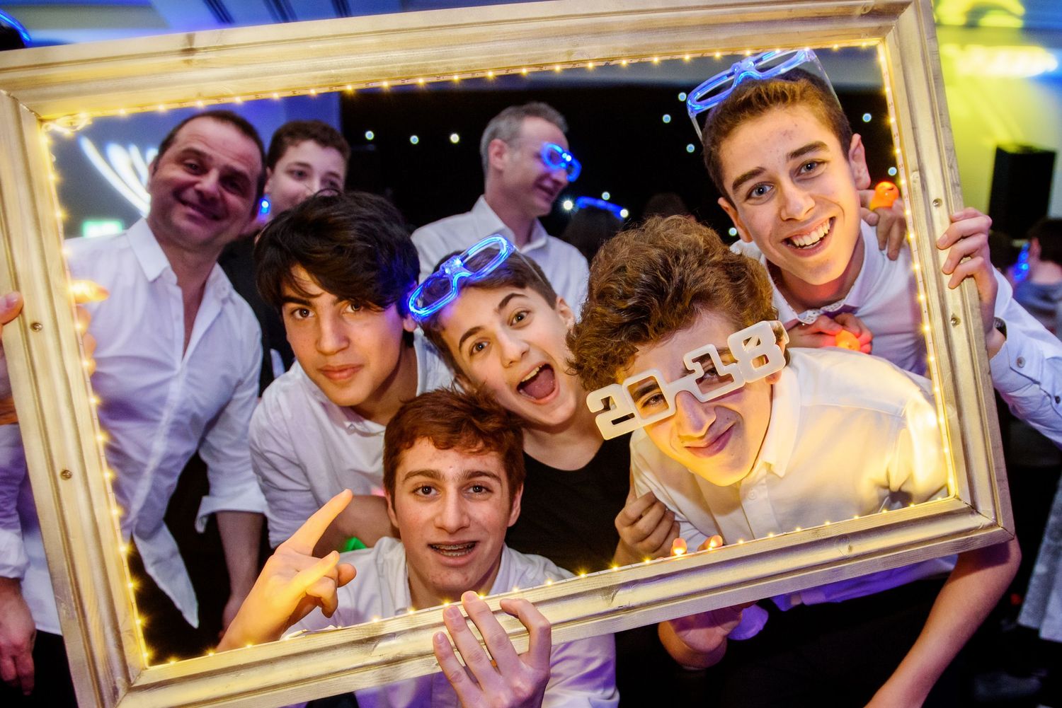 Group of people posing playfully through a decorative gold picture frame at a party with colorful lighting.