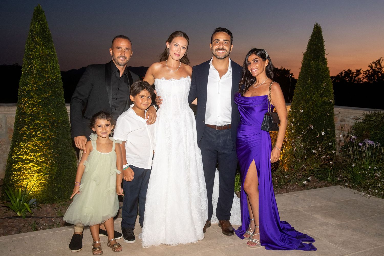 A group of elegantly dressed people pose together at an outdoor evening event with landscaped gardens visible in background.