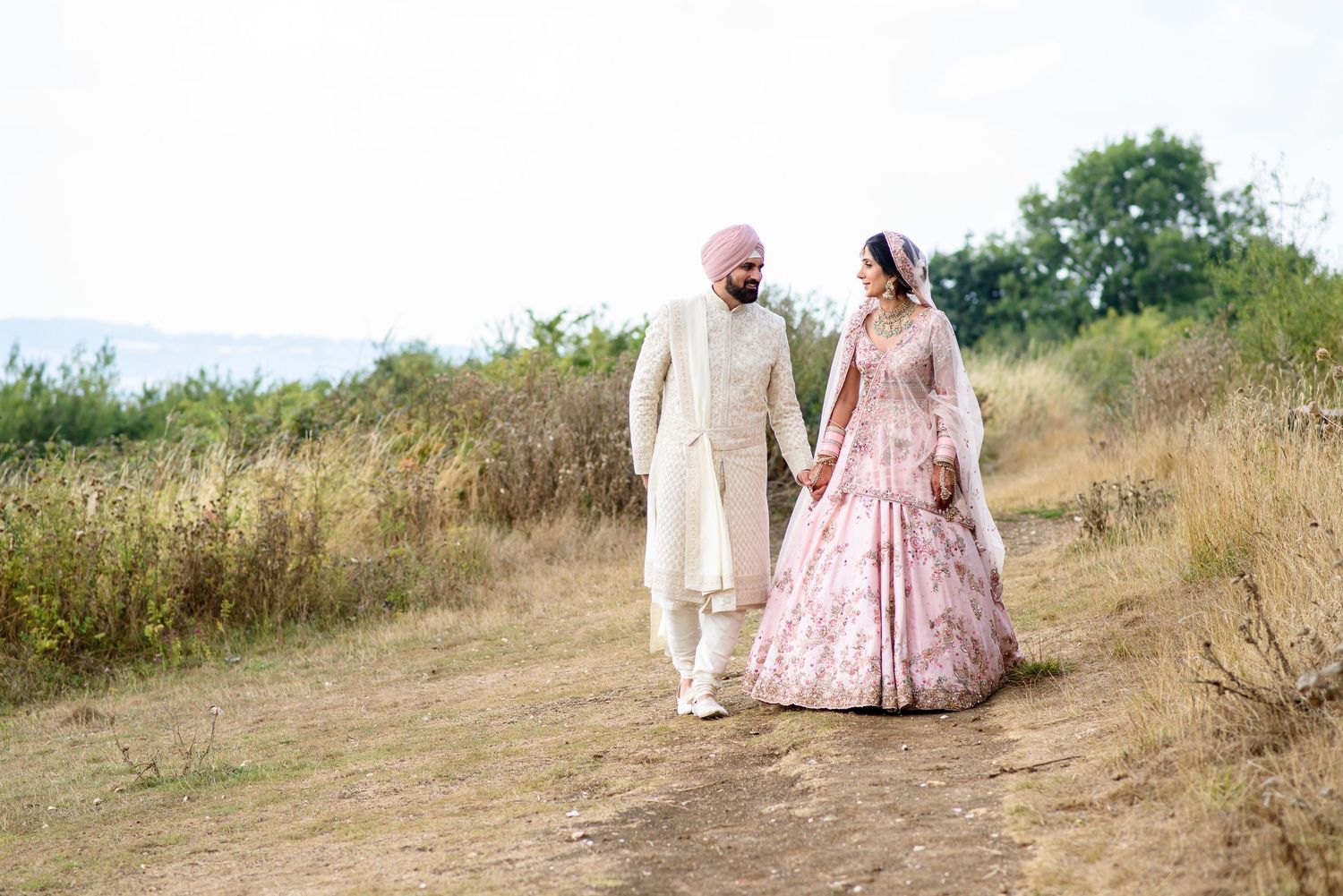 A couple walks together on a dirt path wearing traditional Indian wedding attire in pink and cream colors.
