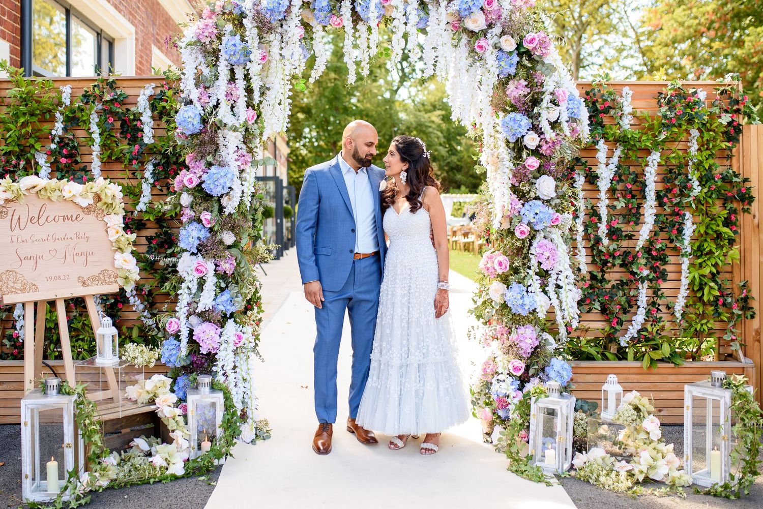 A romantic outdoor wedding ceremony decorated with cascading purple and blue floral arrangements and white fabric draping.