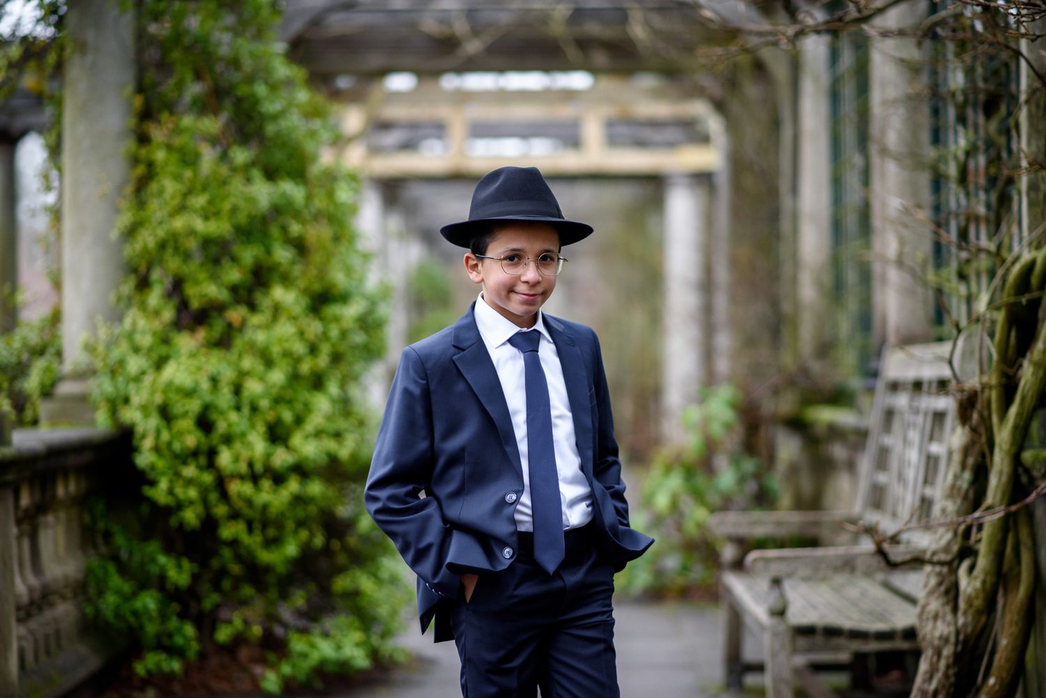 A person in a navy suit and fedora stands in front of stone architecture and greenery.