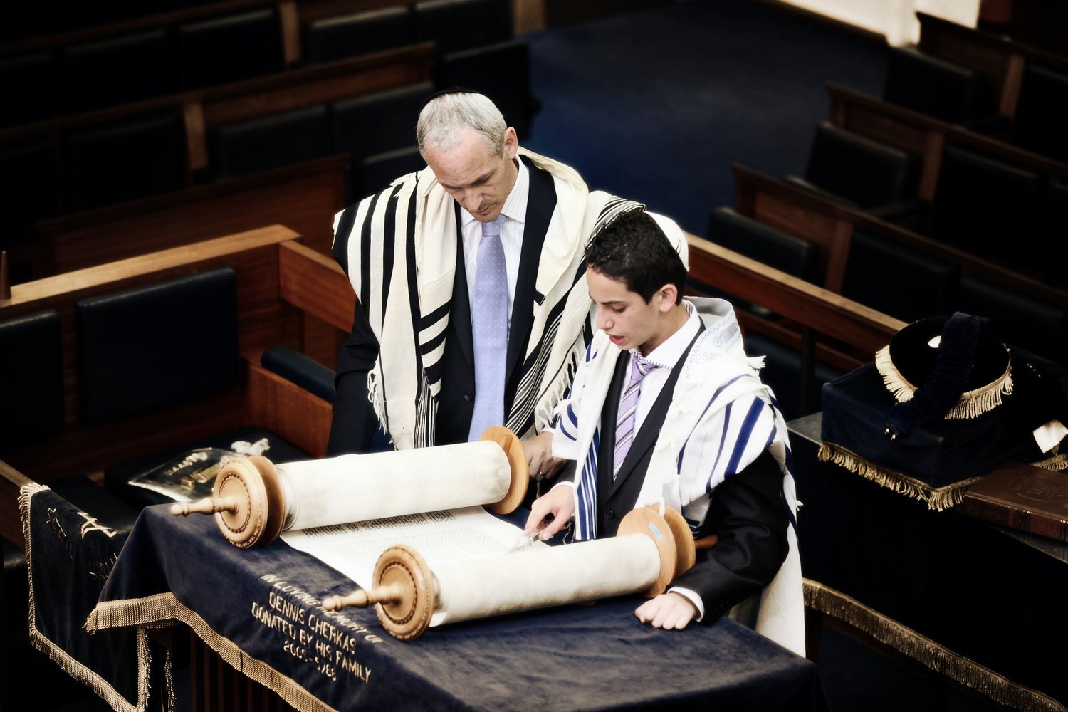 Multiple people in prayer shawls study religious texts at wooden tables.