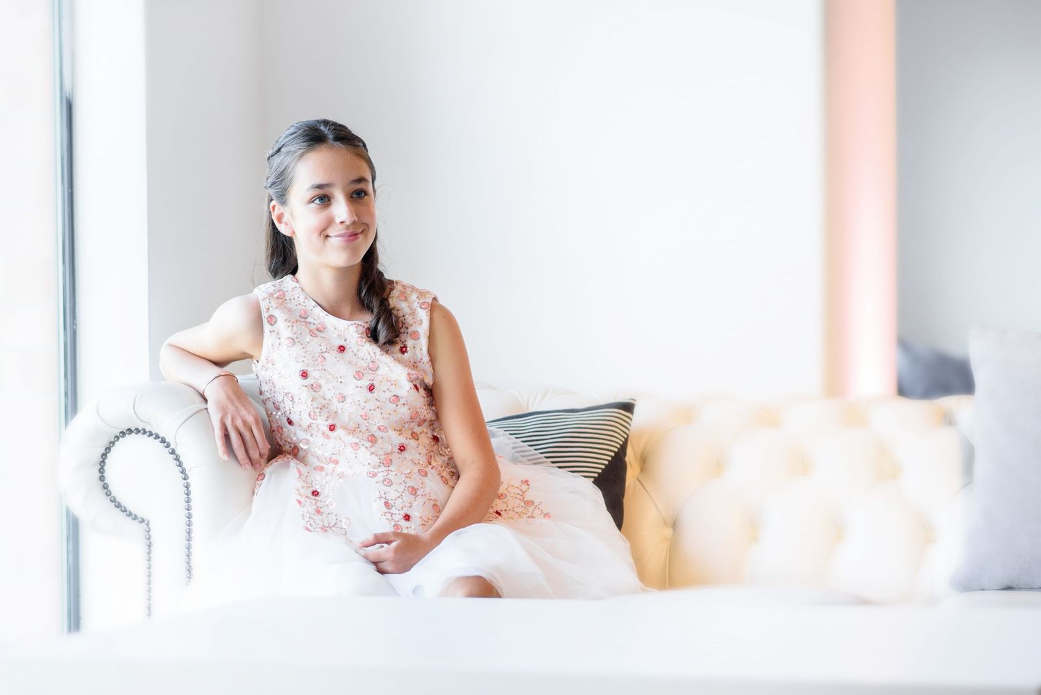 A person in a pink polka dot dress poses on a white bed near bright windows in a sunlit room.