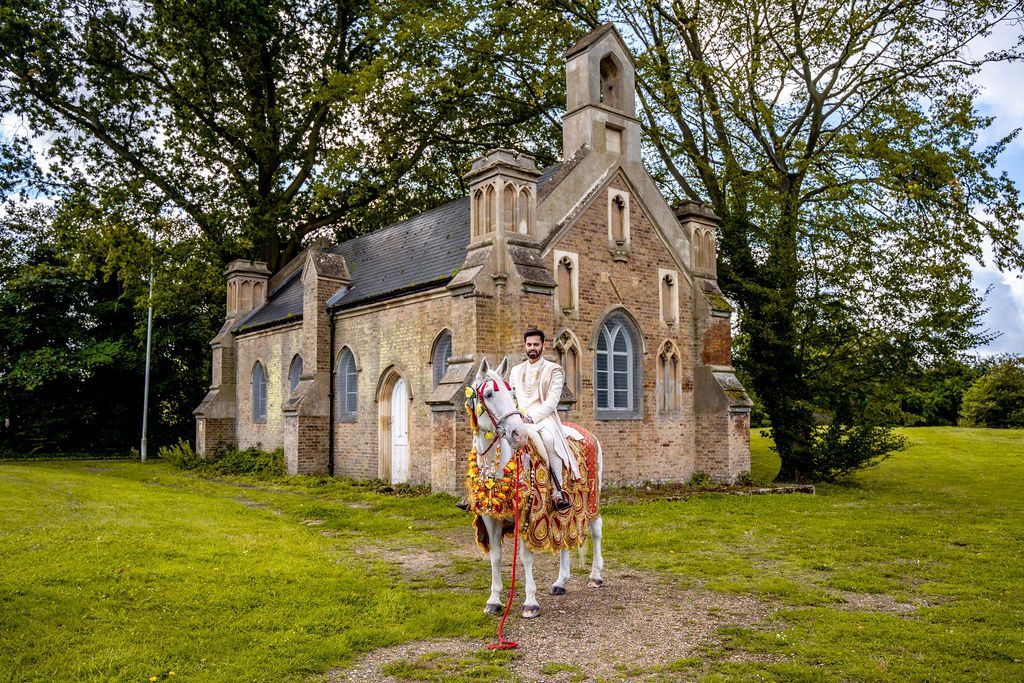 Person in white outfit riding a decorated horse in front of a historic stone chapel surrounded by trees.