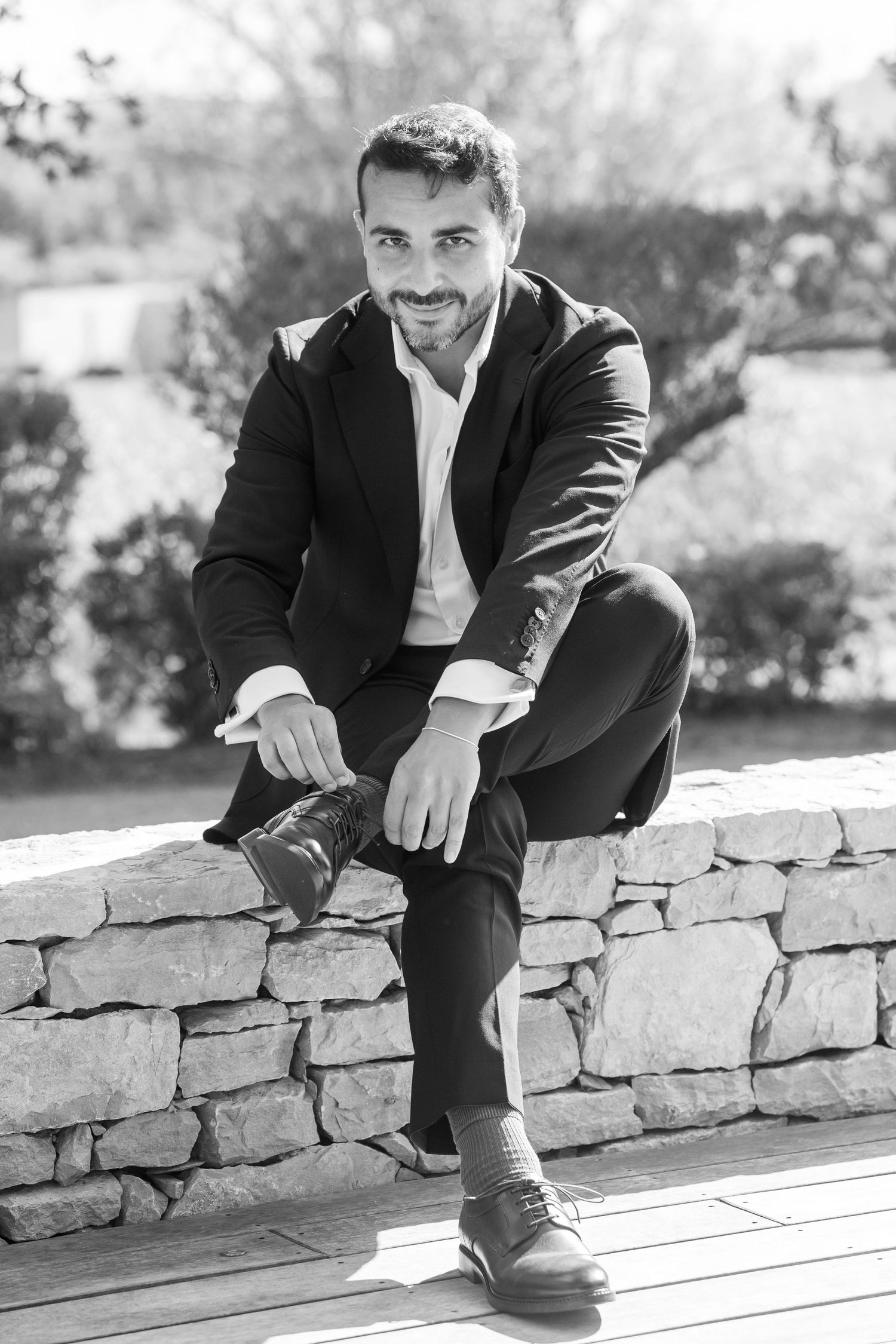A person in a dark suit sits casually on a stone wall in a black and white photo series.