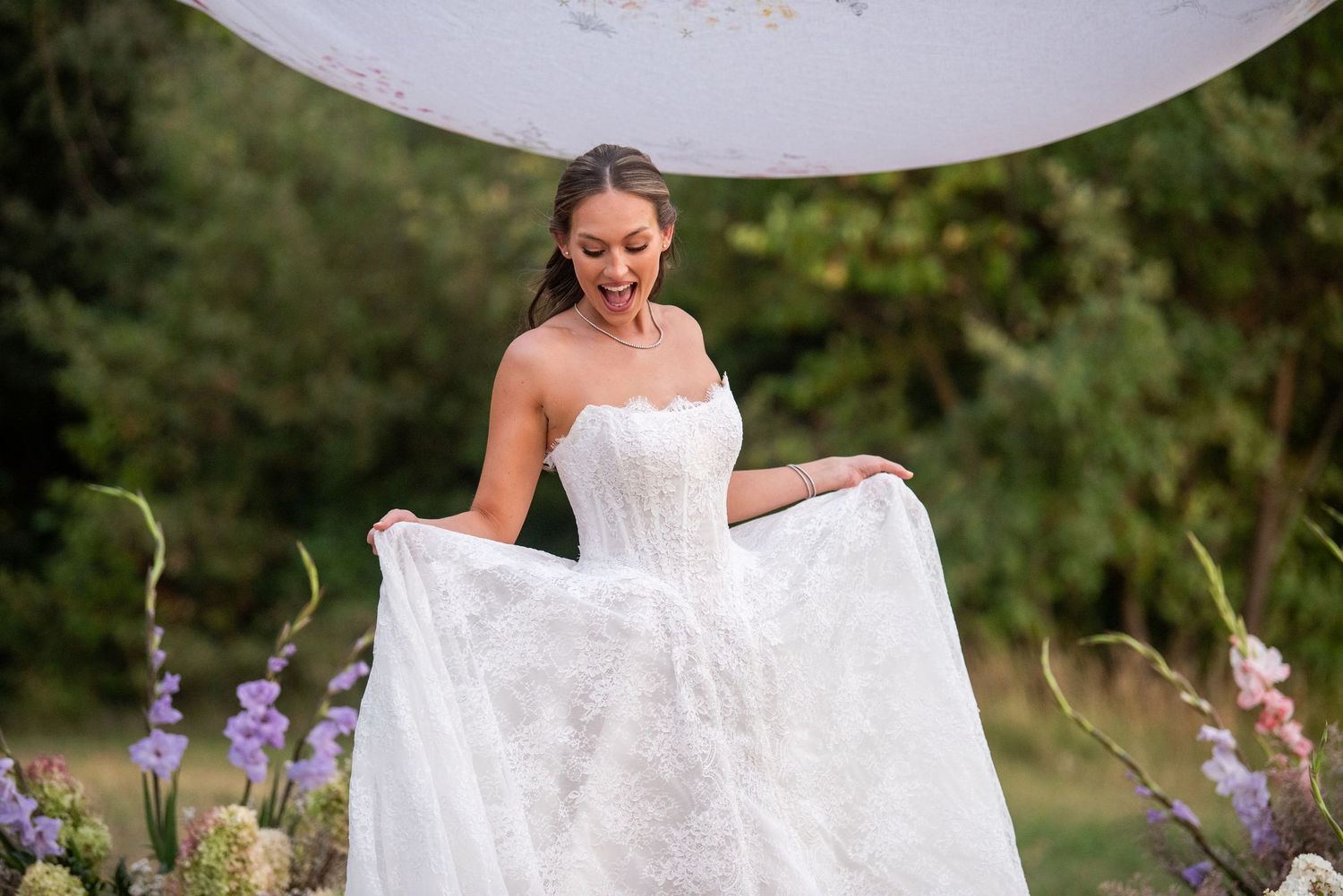 A bride in a white strapless wedding gown stands outdoors among purple wildflowers on a sunny day.