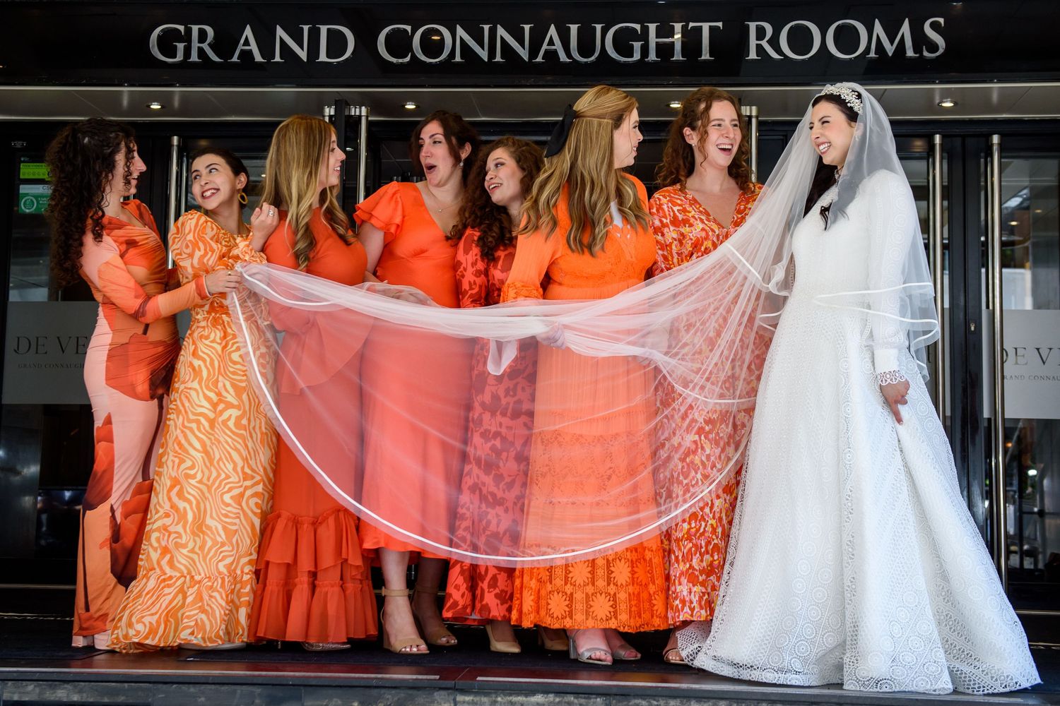 Wedding party in orange bridesmaid dresses poses in front of the Grand Connaught Rooms sign.
