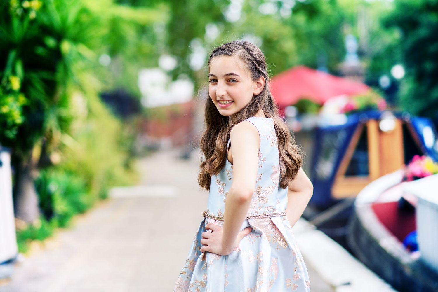 A person in a white dress poses outdoors in a garden setting with lush greenery and flowers in the background.