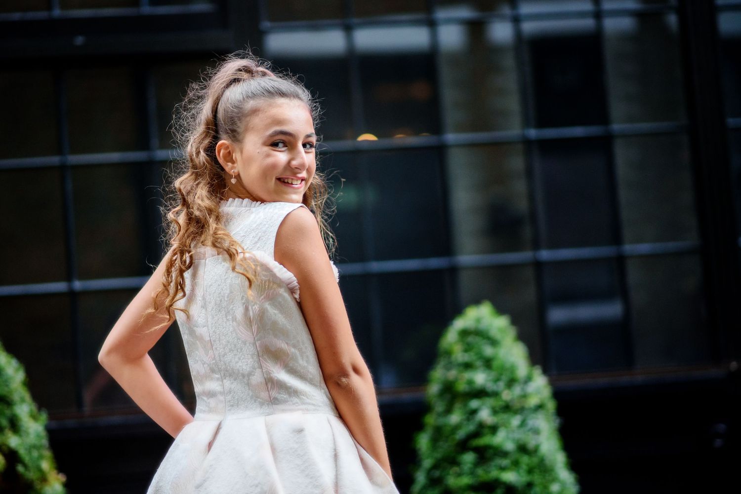 A bride in a white lace dress twirls and dances outdoors with greenery and buildings in the background.