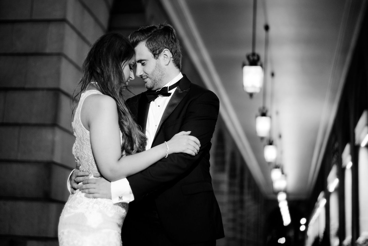 Romantic black and white photo of a couple dancing in formal attire under illuminated archways.