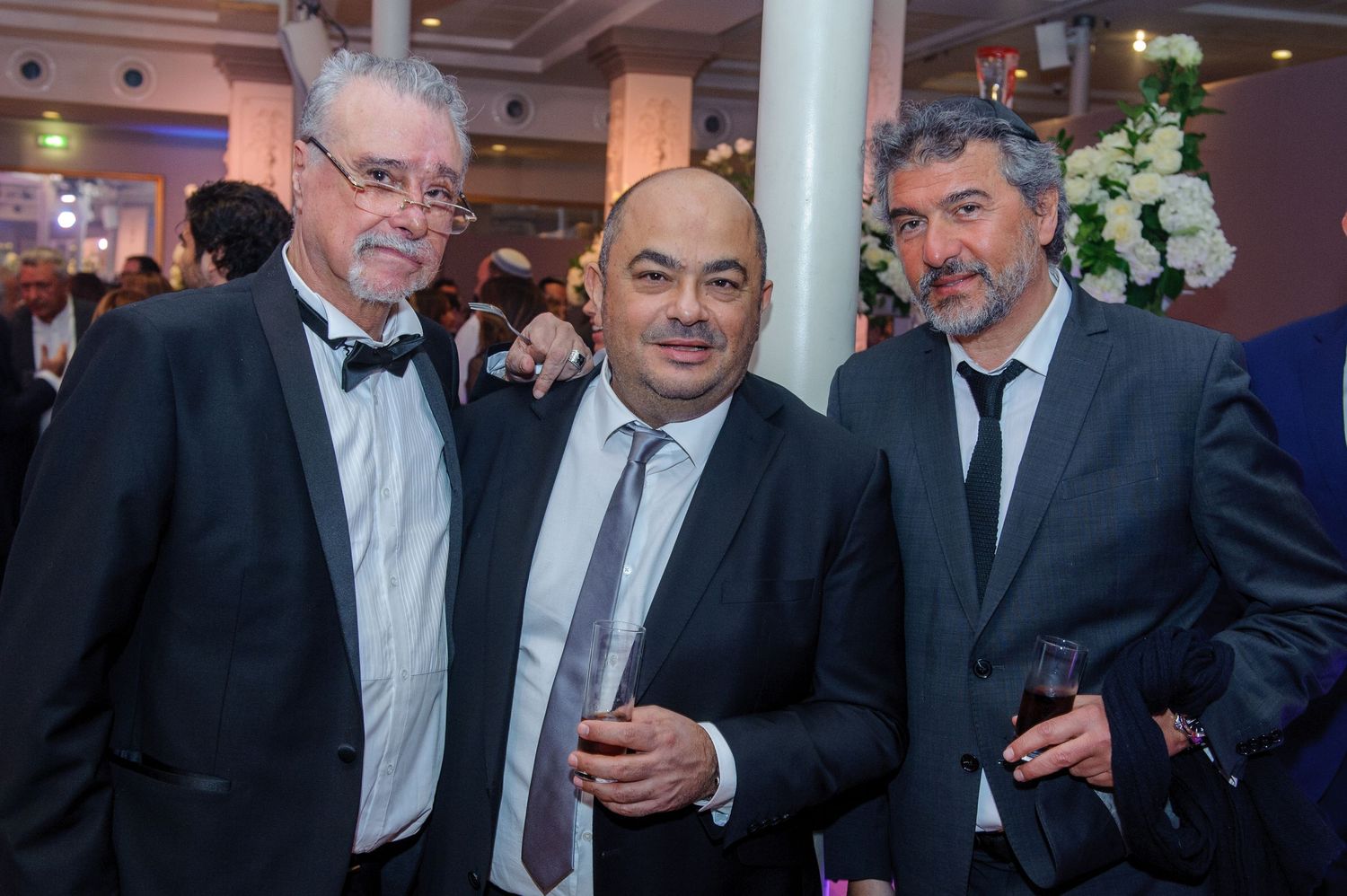 Three men in formal attire having a conversation at an indoor event with decorative lighting and white flowers.