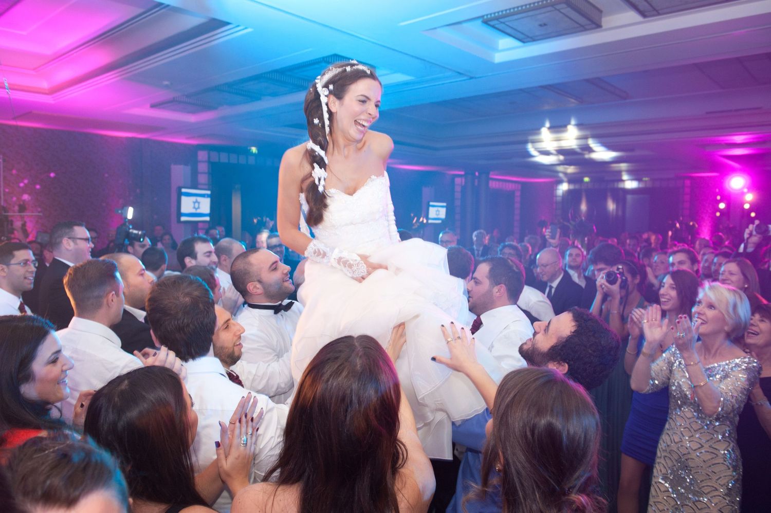 Wedding guests dance and celebrate on a dance floor illuminated by vibrant pink and blue lighting at a reception party.