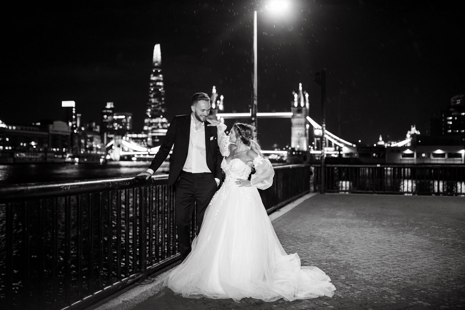 Black and white wedding photo of bride and groom by Thames with Tower Bridge and The Shard lit up at night.