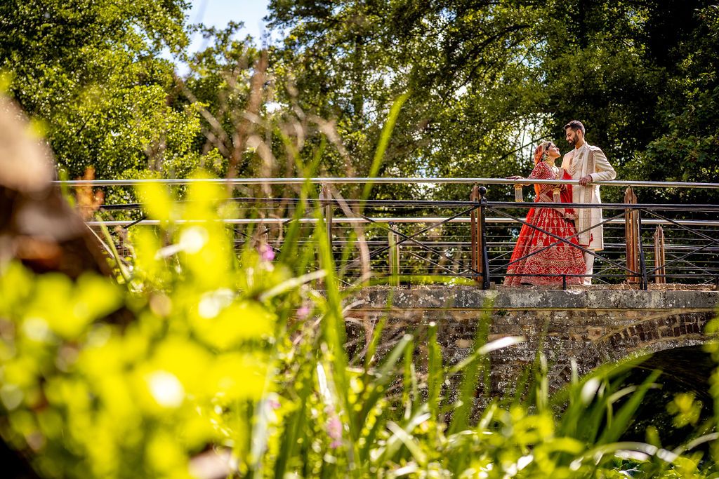 Couple in traditional attire standing on a rustic bridge surrounded by lush green trees and foliage.