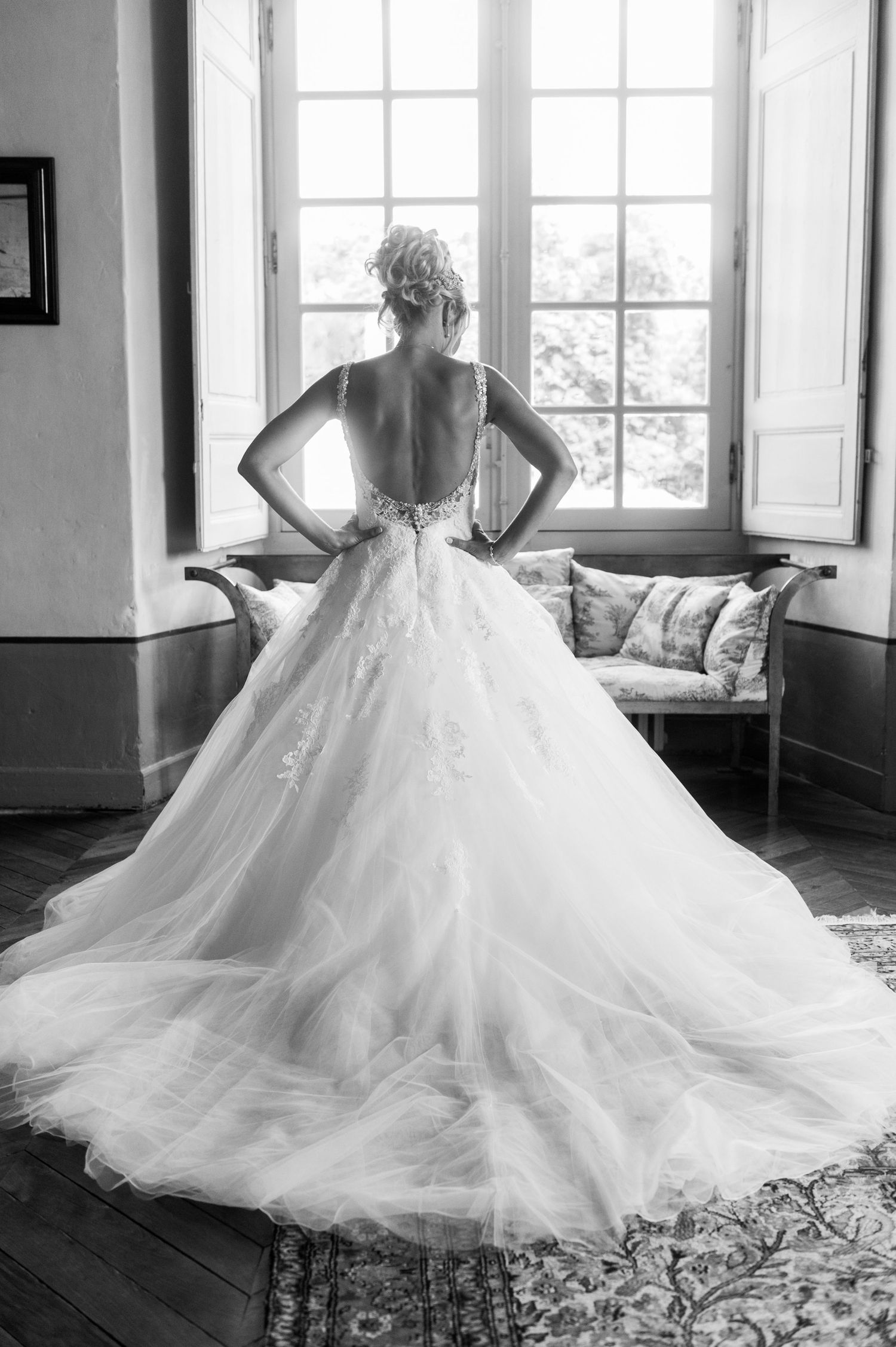 Black and white photo of a bride in a stunning backless ball gown with a long train, standing before a window.