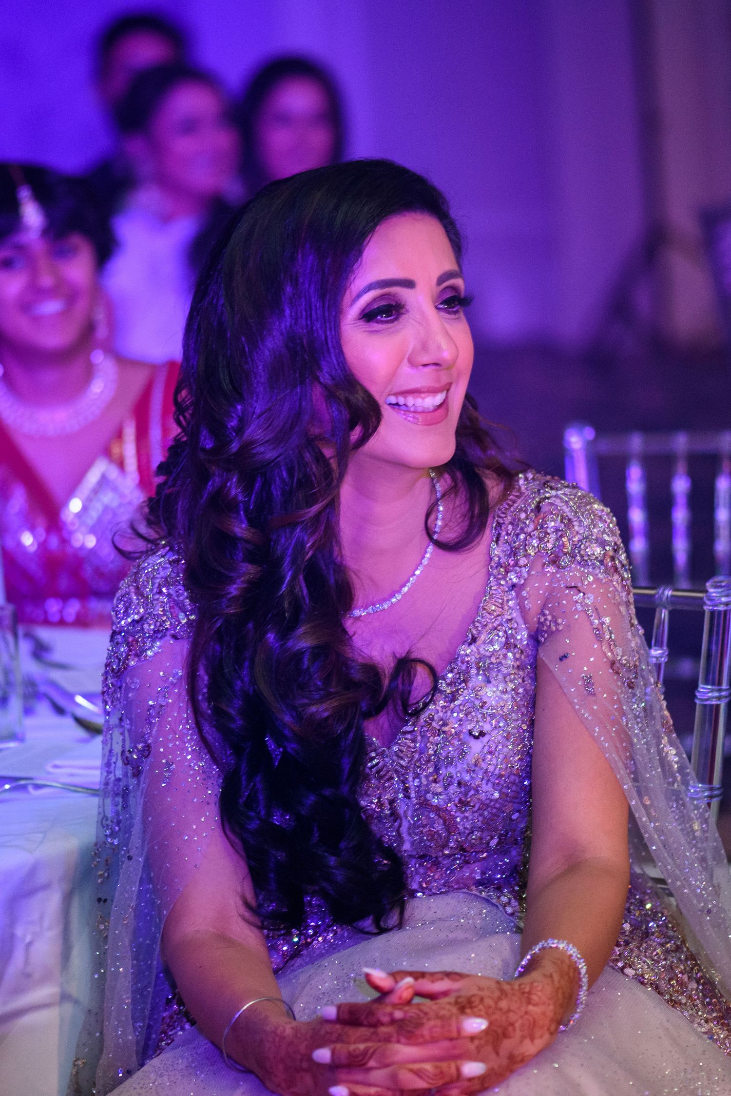 A woman in a sparkly evening gown sits at a decorated reception table under purple lighting.
