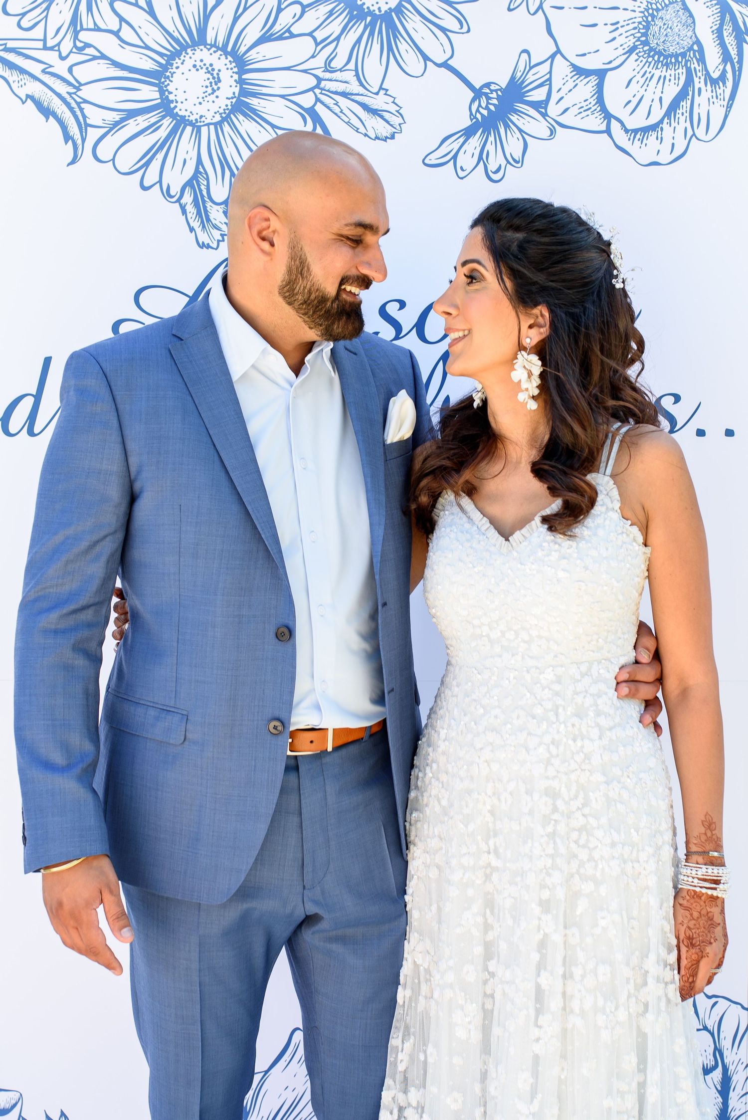 Newlywed couple poses against floral backdrop in blue suit and white lace dress during wedding photoshoot.