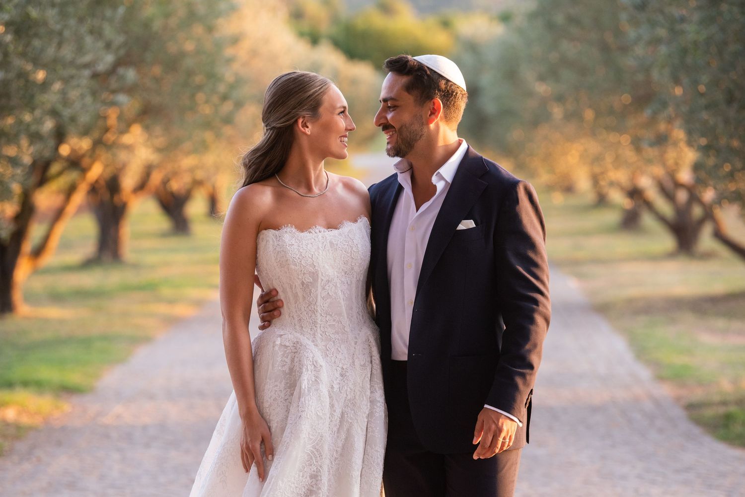 A couple in wedding attire walks together through an olive grove at sunset during their romantic photoshoot.