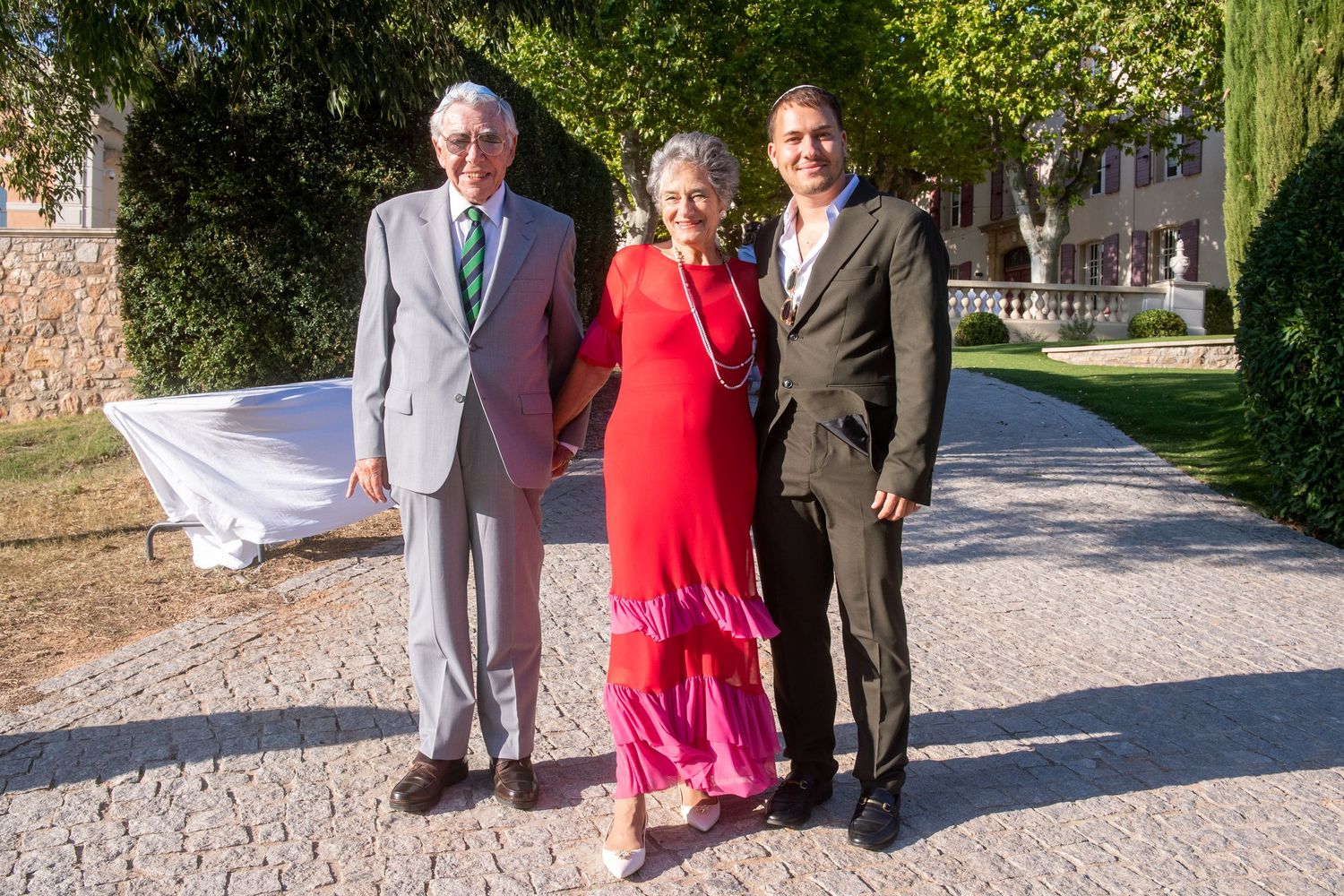 Three people pose together outdoors on a sunny day, with a woman in a flowing red dress standing between two men in suits.
