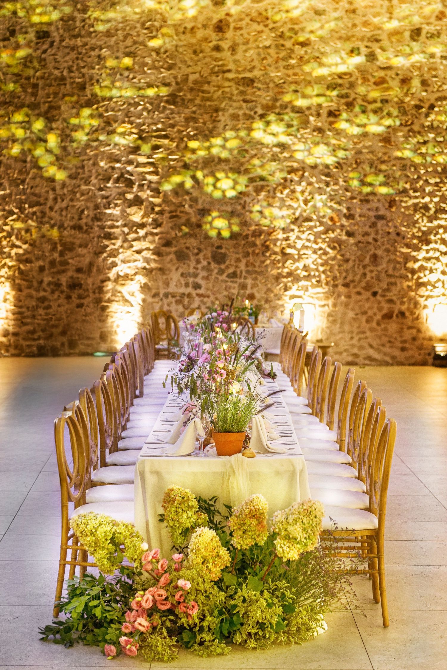 Elegant wedding reception tables with wooden chairs and lush floral centerpieces arranged against a rustic stone wall.