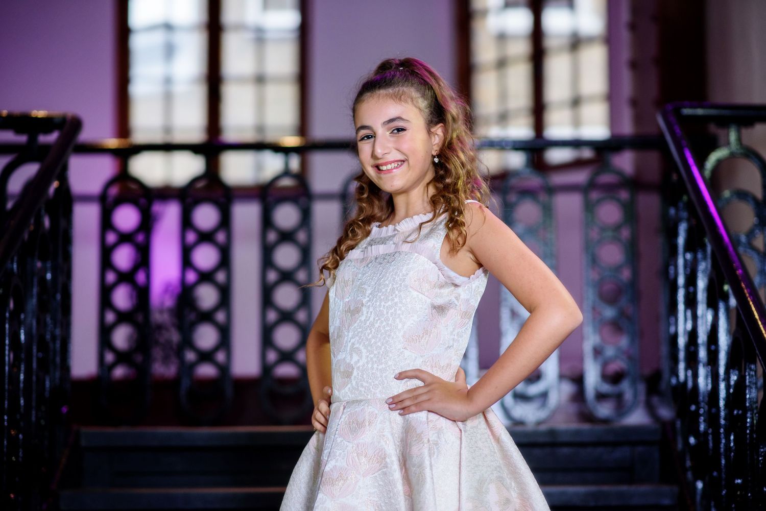 A bride in a white lace wedding dress poses elegantly in front of ornate windows with purple lighting.
