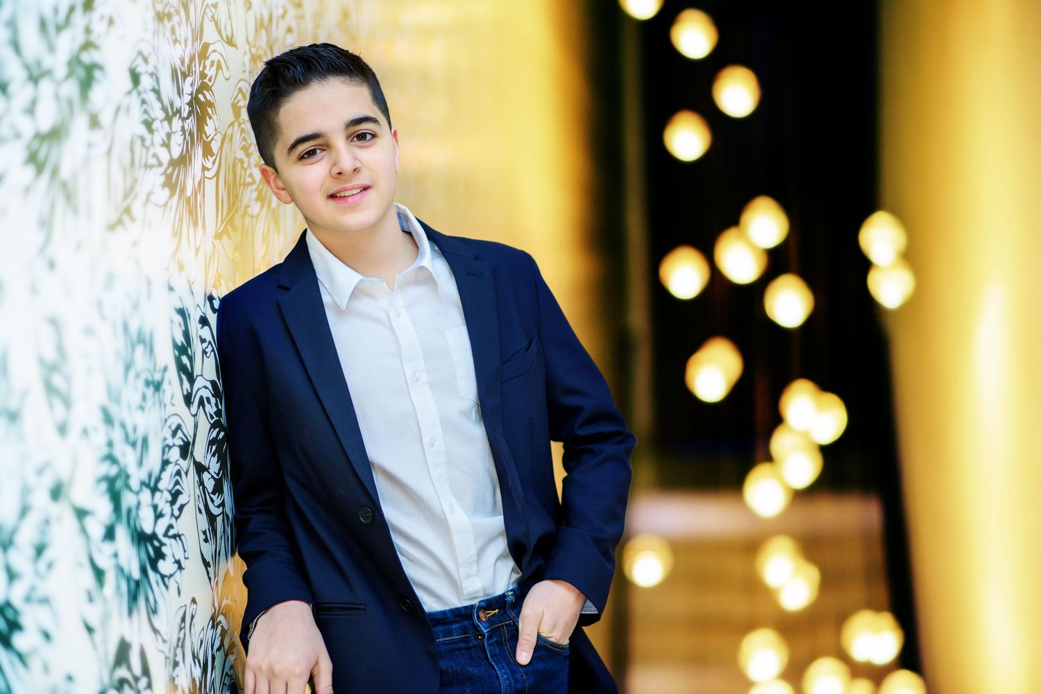Person in navy suit and white shirt poses in front of warm glowing lights and greenery for formal portrait.