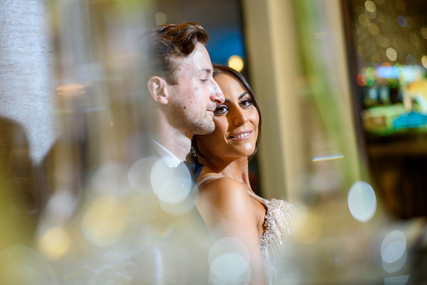 A bride and groom share a romantic moment, captured through a soft bokeh foreground at an elegant indoor venue.