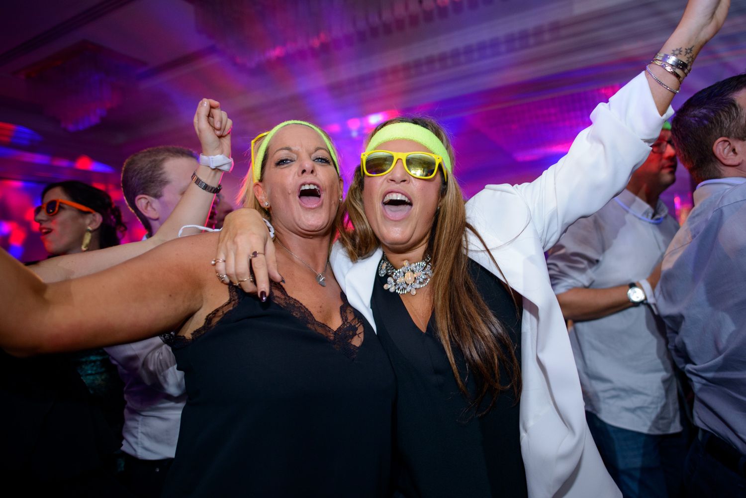 Partygoers wearing colorful sunglasses celebrate enthusiastically on a dance floor with purple lighting.