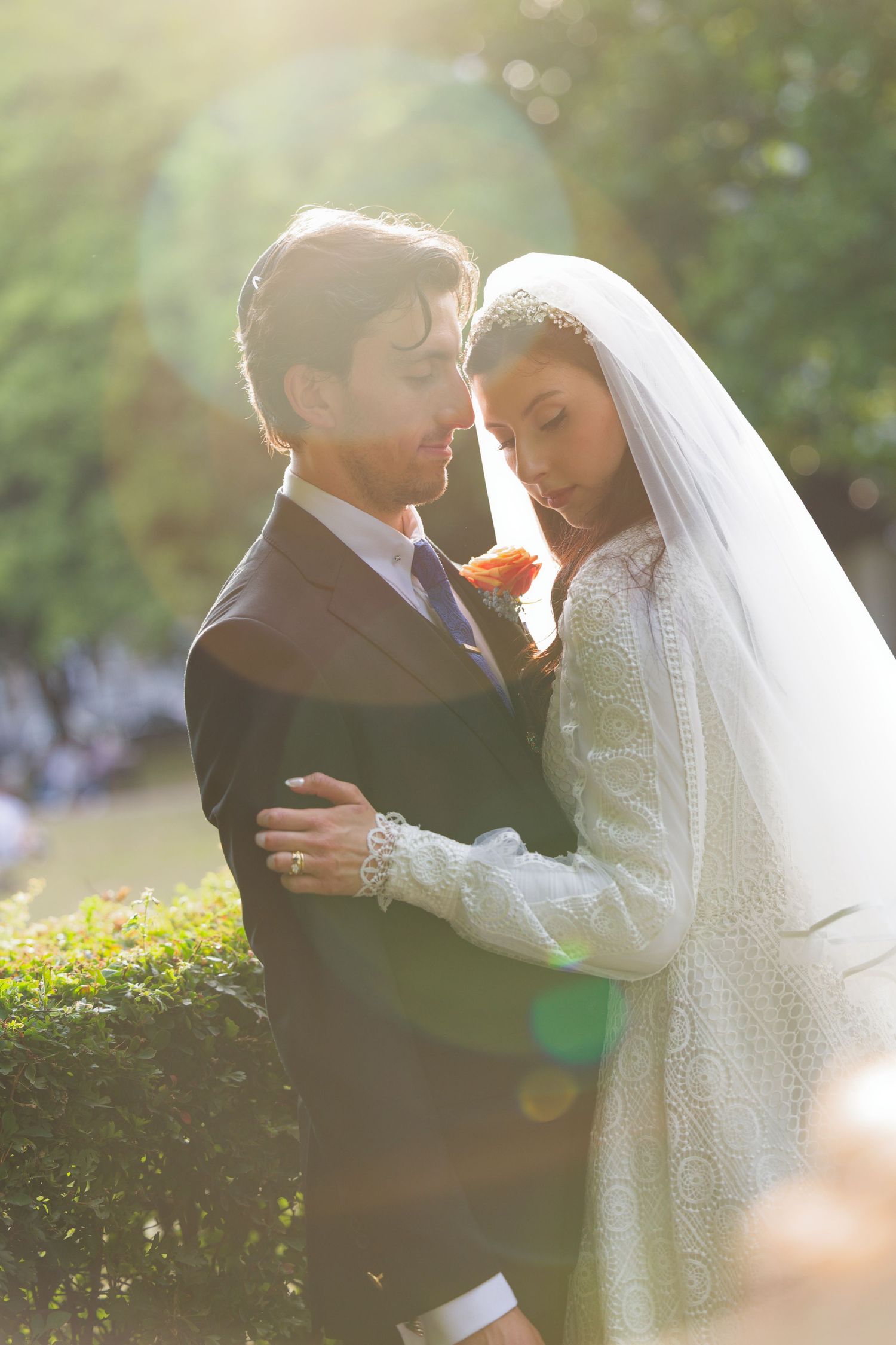 A romantic wedding couple embraces outdoors in a garden setting with soft sunlight filtering through the trees.