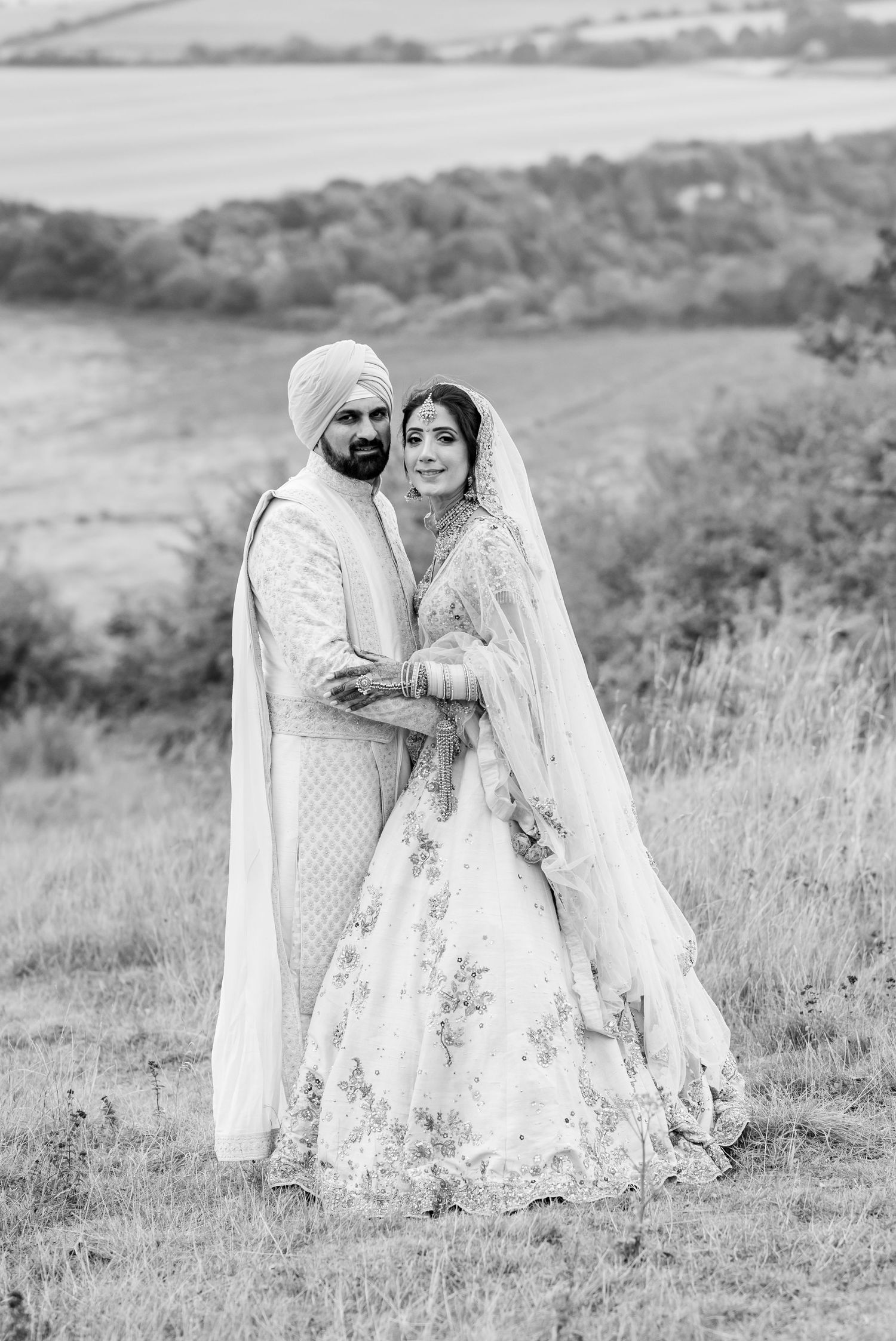 A couple in traditional Indian wedding attire poses together in a scenic meadow with rolling hills in black and white.