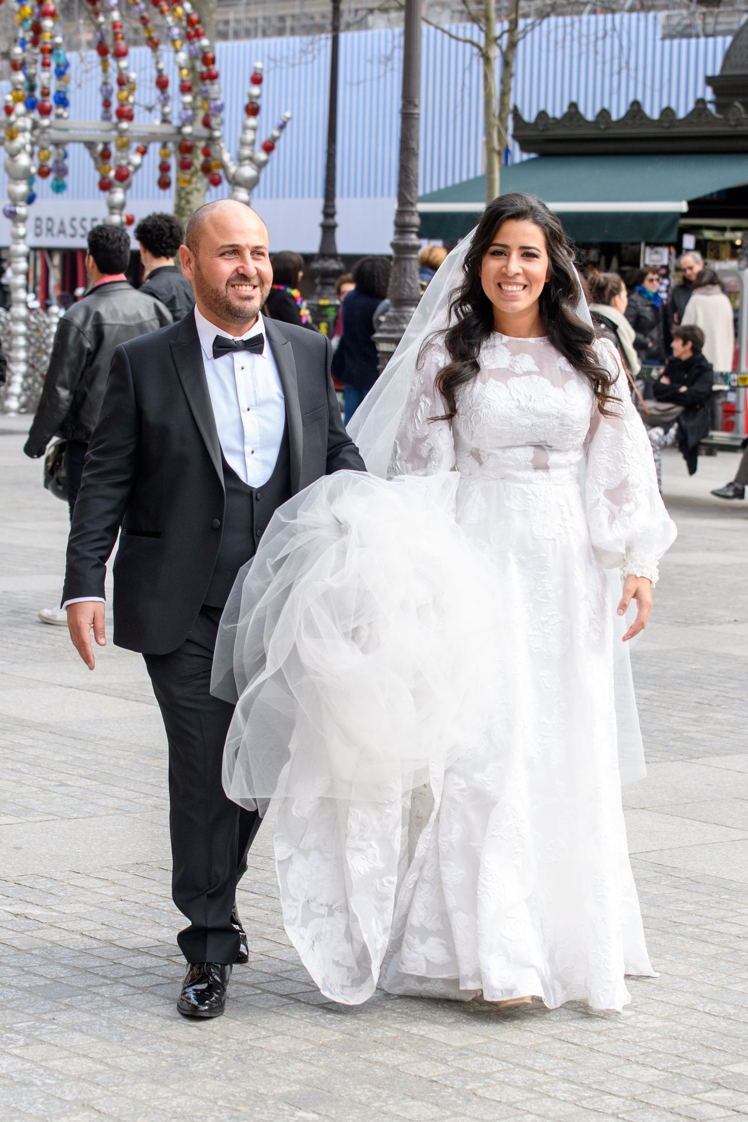 A couple in wedding attire walks together outdoors under decorative red lanterns and traditional Chinese decor.