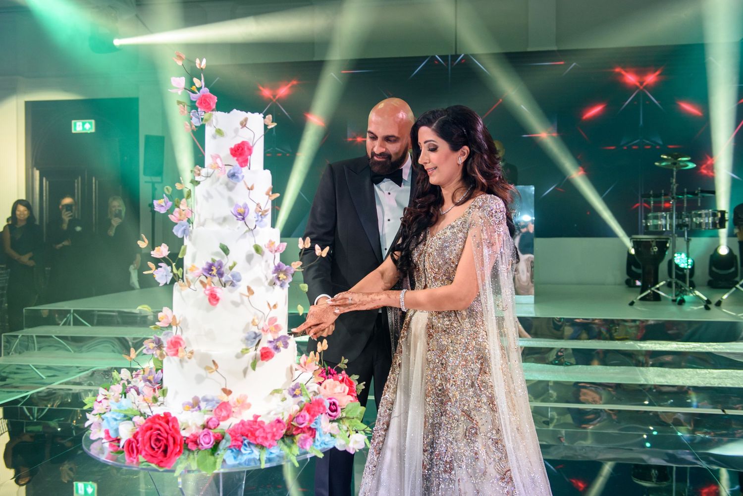 Couple cuts elegant white wedding cake with pink floral decorations under dramatic green lighting at reception.