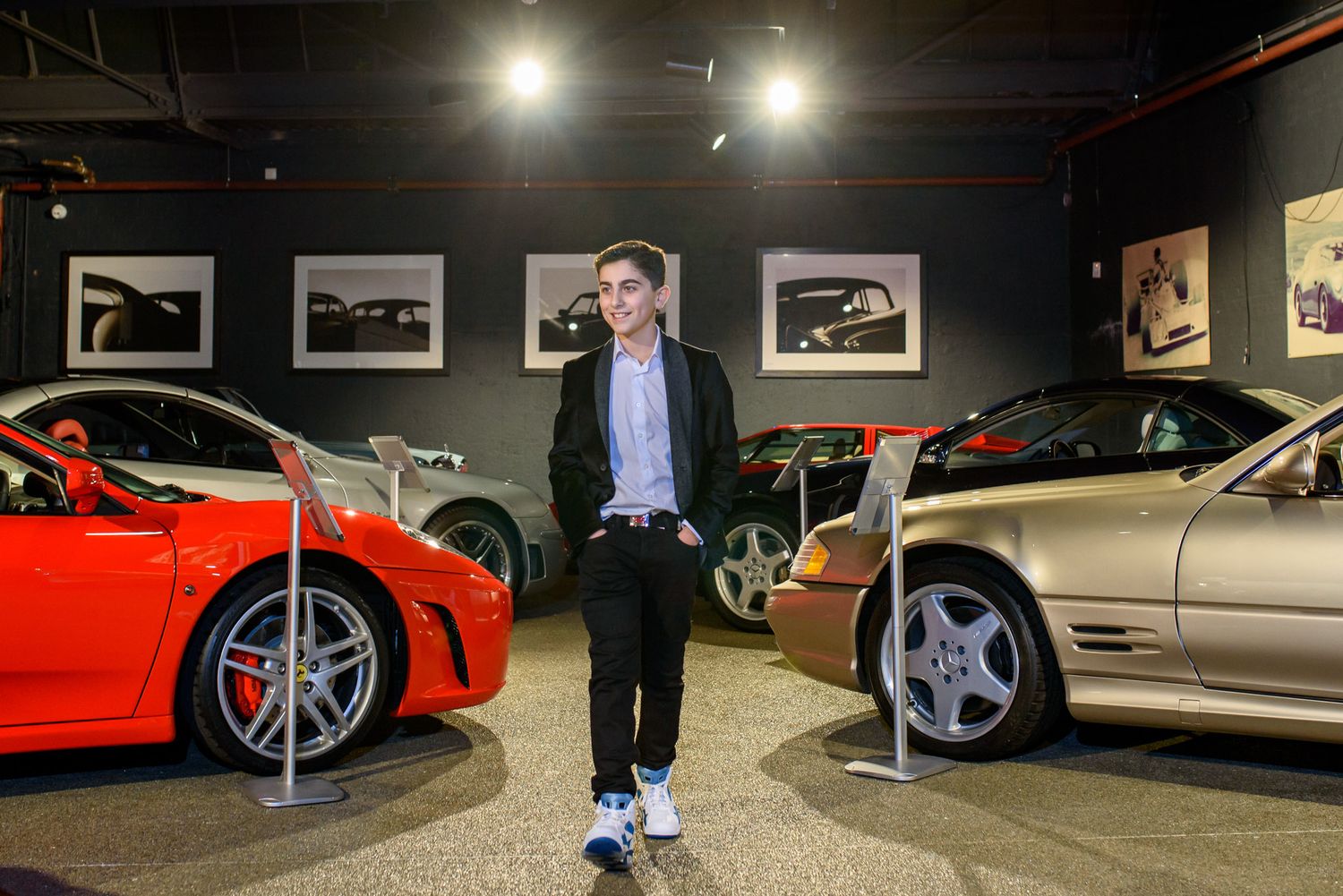A dark car showroom displays multiple luxury vehicles under bright spotlights with framed artwork on the walls.