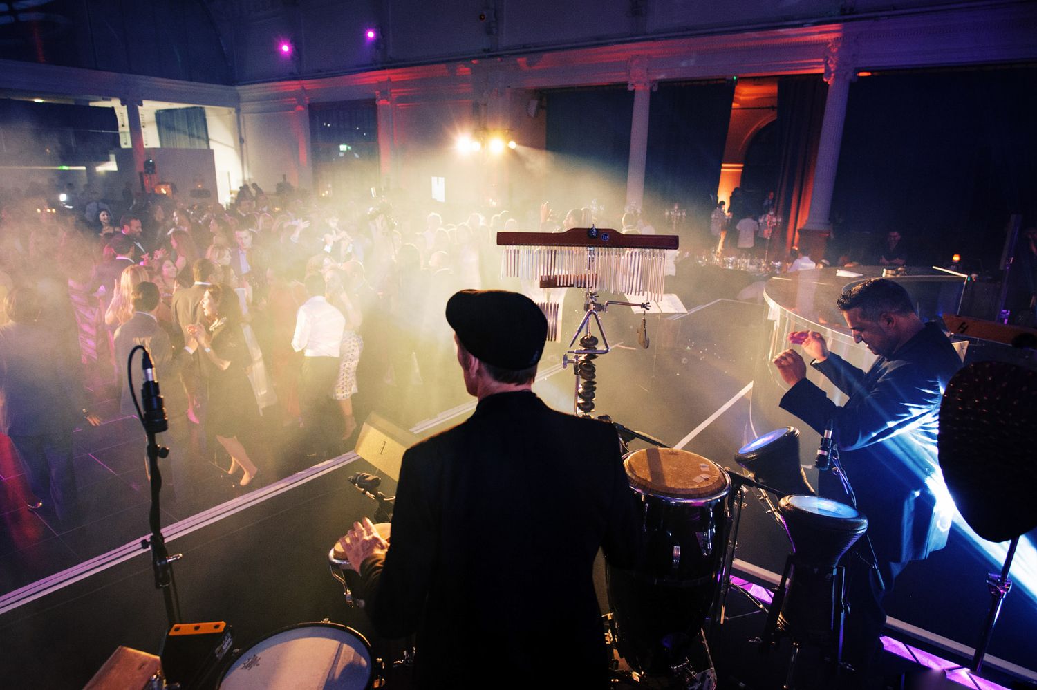 A dark concert venue with hazy atmosphere illuminated by colorful stage lights and silhouettes of performers on stage.