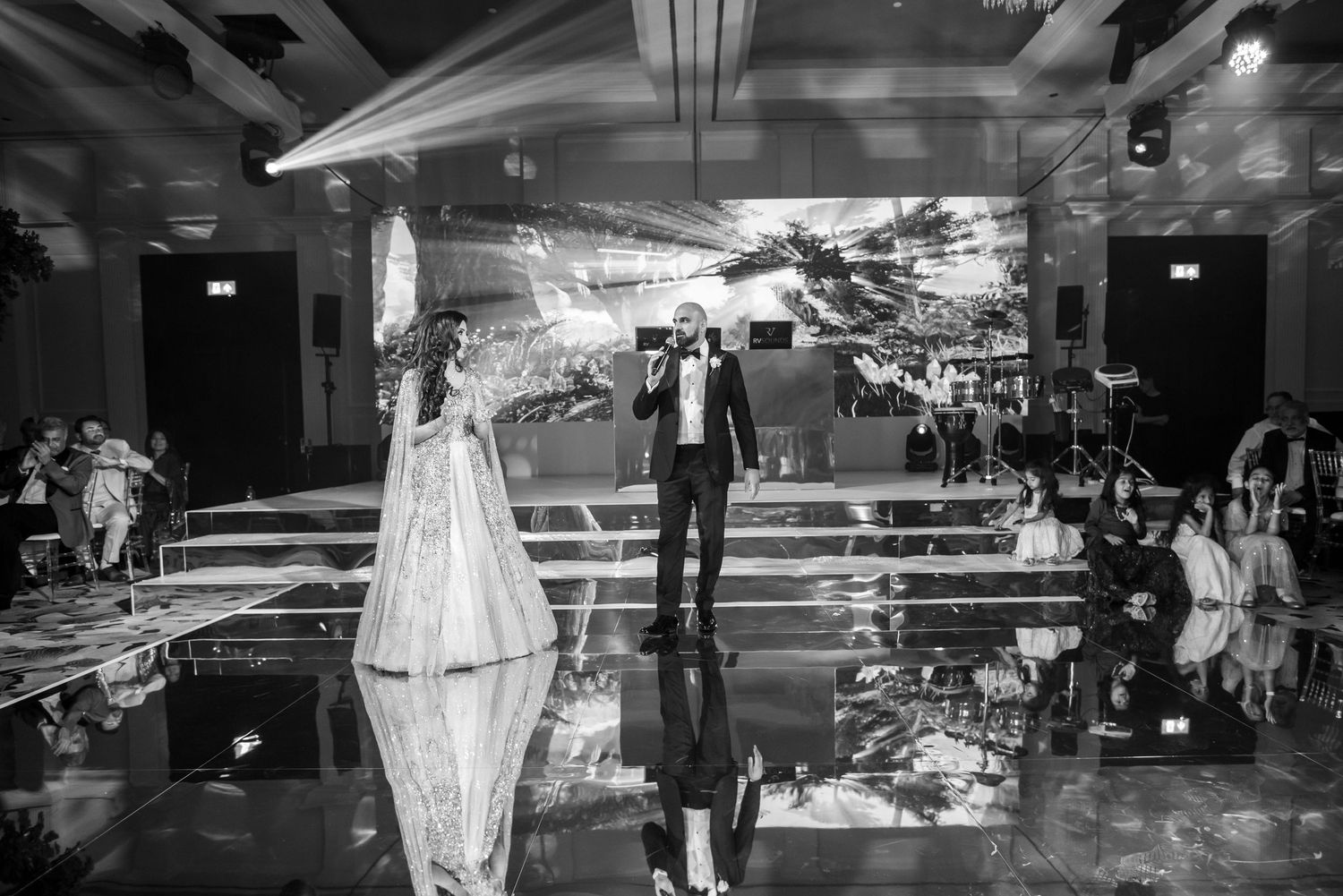 A couple performs their first dance at a wedding reception on a reflective black dance floor with city backdrop.