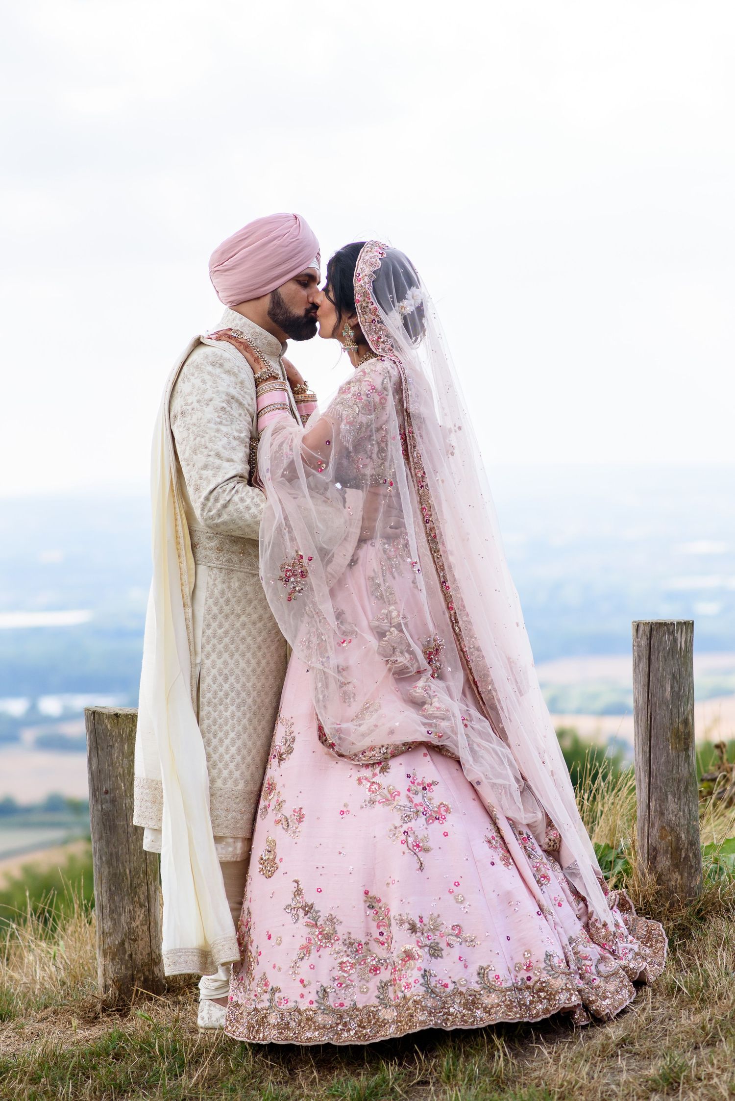 A couple in traditional Sikh wedding attire share an intimate moment outdoors against a scenic mountain backdrop.