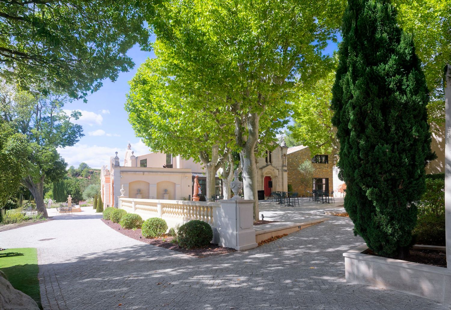 Mediterranean villa with covered terrace and stone driveway surrounded by lush green trees and cypress.