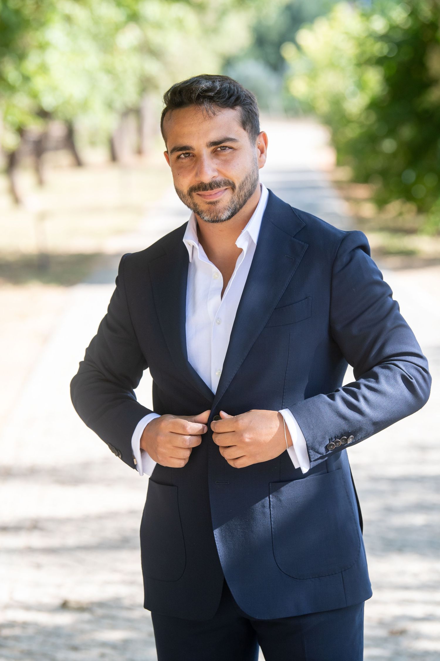 Confident man in navy blue suit buttoning jacket outdoors in tree-lined park path, smiling at camera.