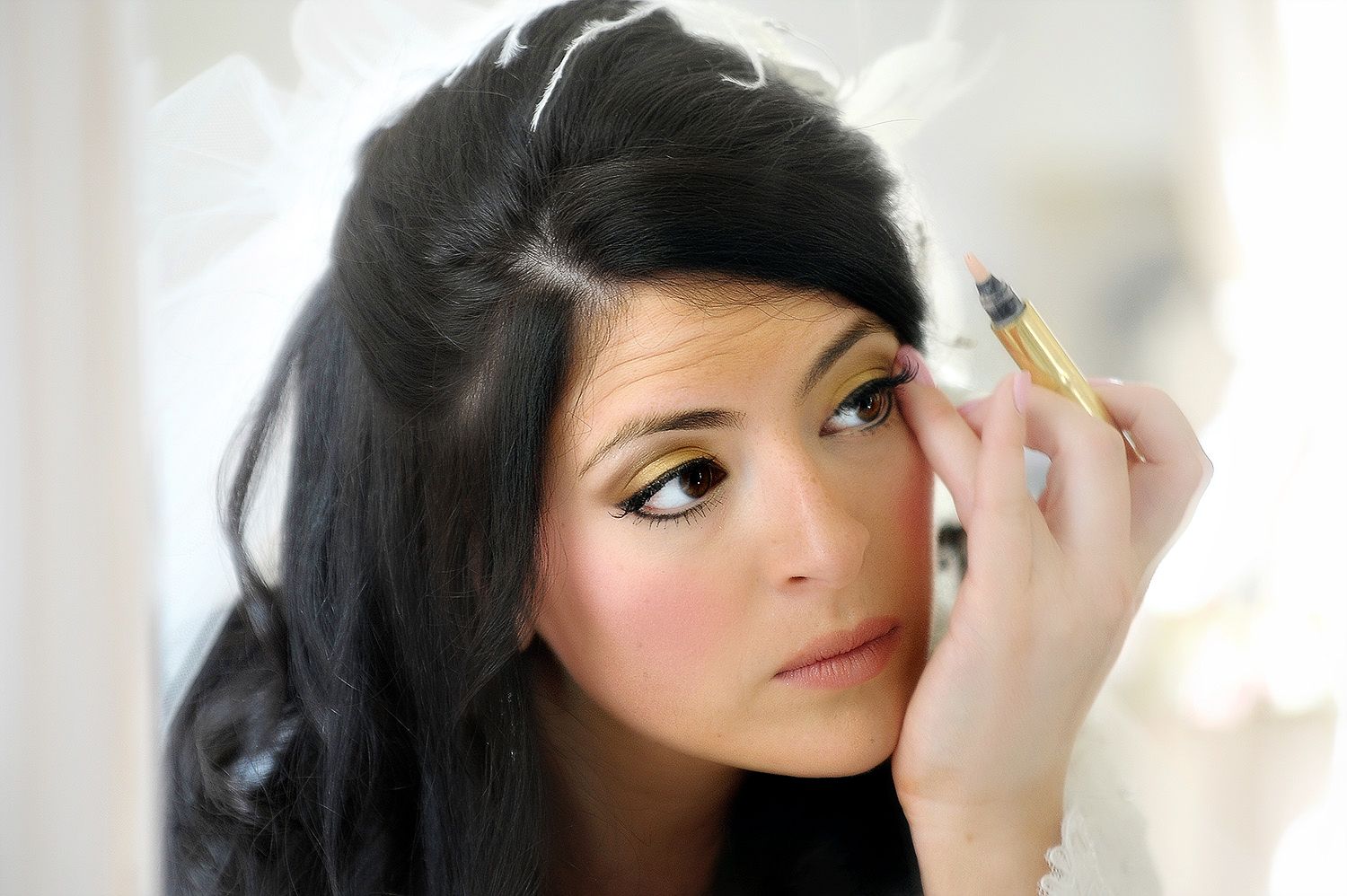 Young woman with dark hair applying eye makeup with a gold pencil, showcasing a glamorous beauty look.