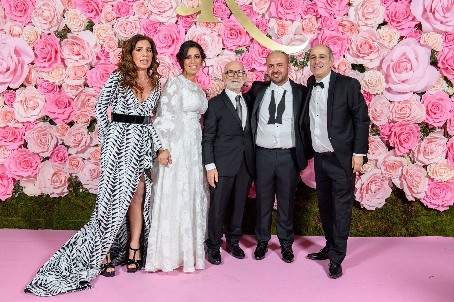 A group of five people pose together in formal attire against a pink floral rose wall backdrop on a pink carpet.